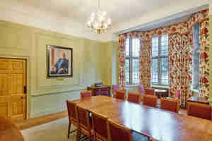 A whole room shot of the Atlantic Room in Rhodes House. This angle looks diagonally down from the head of a long wooden table. On the left wall is a large wooden door and a portrait of a gentleman sat in a chair.  In the wall to the right of this is a large bay window framed by floral curtains.