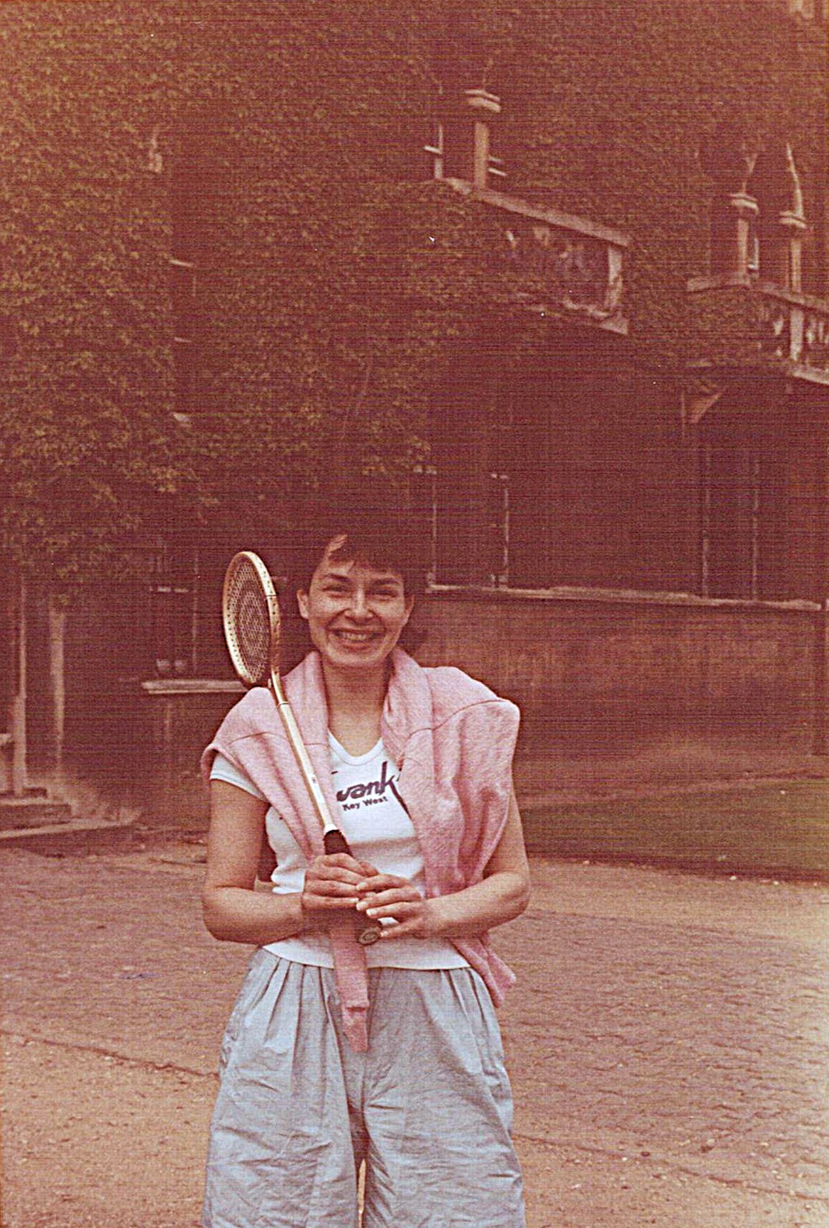 Susan stood before a college building smiling and holding a squash racket. She is wearing athleisure.
