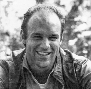 A black-and-white headshot of Peter. He's wearing a thick shirt over a t-shirt. He is smiling and behind him are trees. 