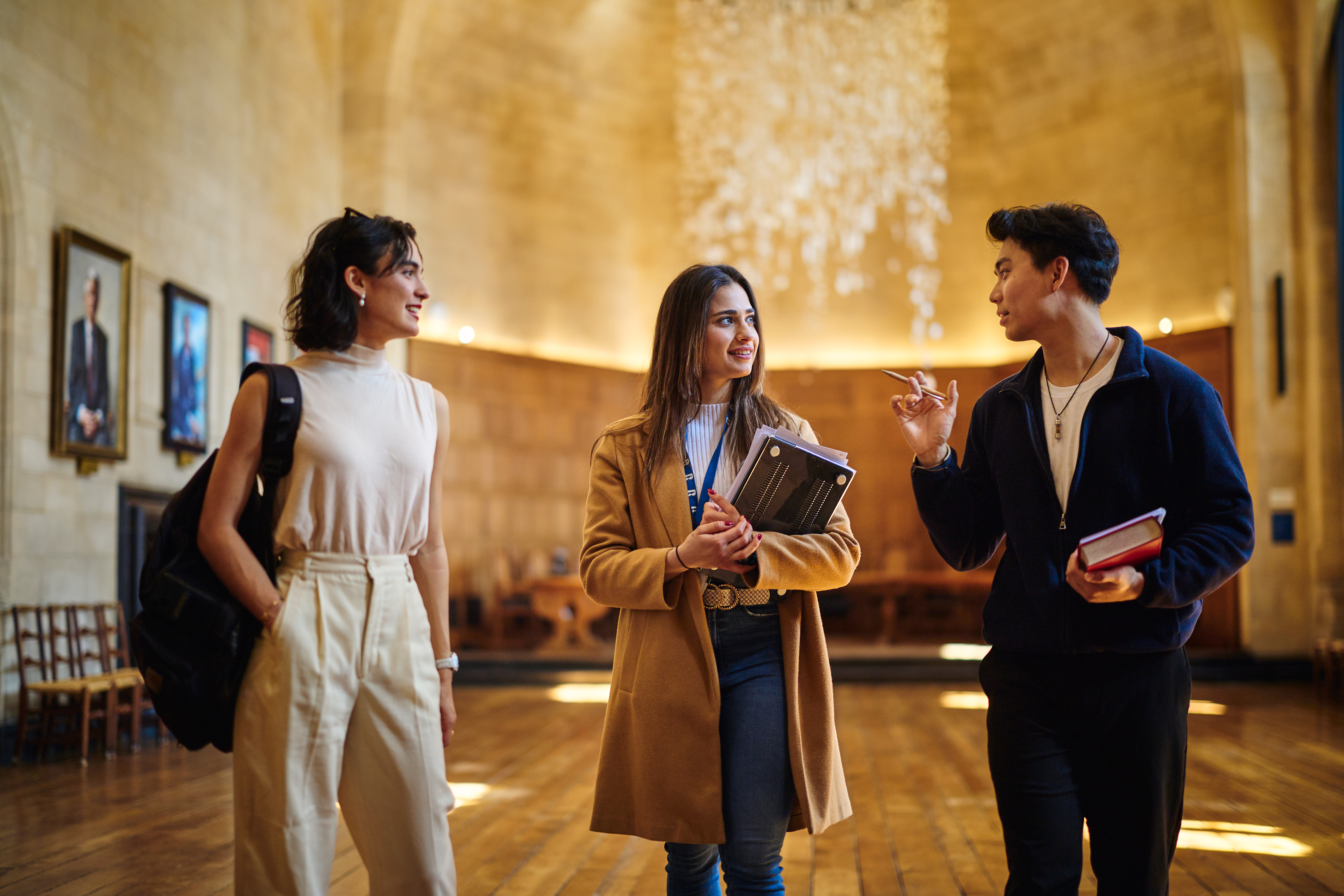 Three students engaged in a lively discussion. They are standing in a large hall with wood panelling with portraits on the walls and a sculpture descending from the ceiling in the background, and they are holding books and backpacks.