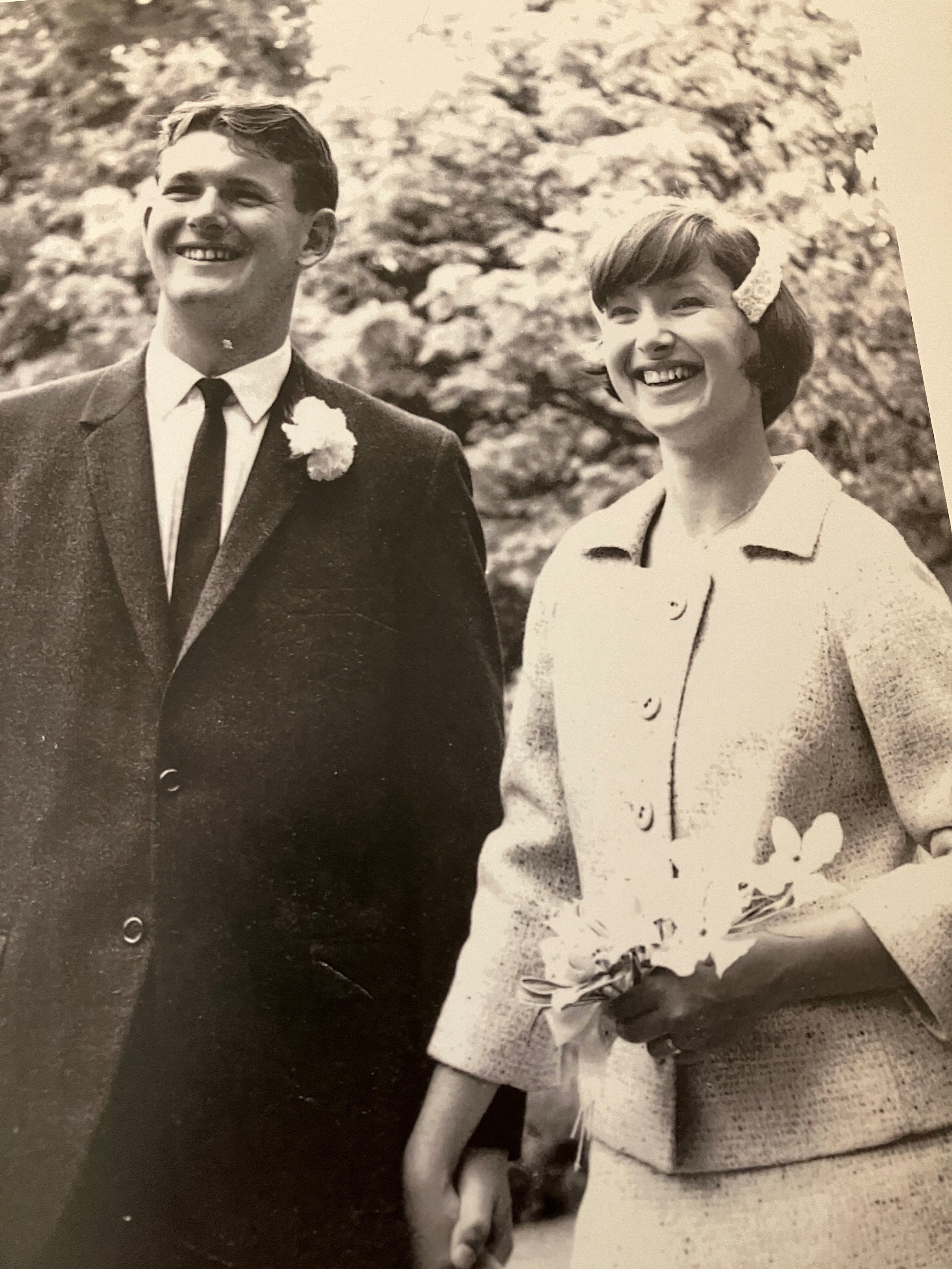 A black-and-white picture of Nick standing with his wife. Nick is in a suit and his wife is wearing a white dress. There are trees in the background.