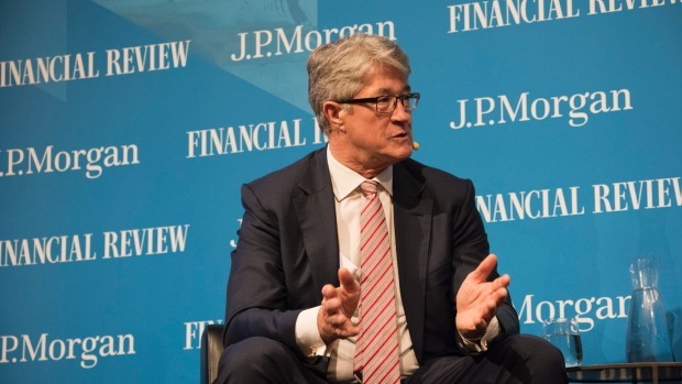 Mike is sat in a suit speaking. Behind him is a blue wall which lists sponsors such as 'J.P. Morgan'