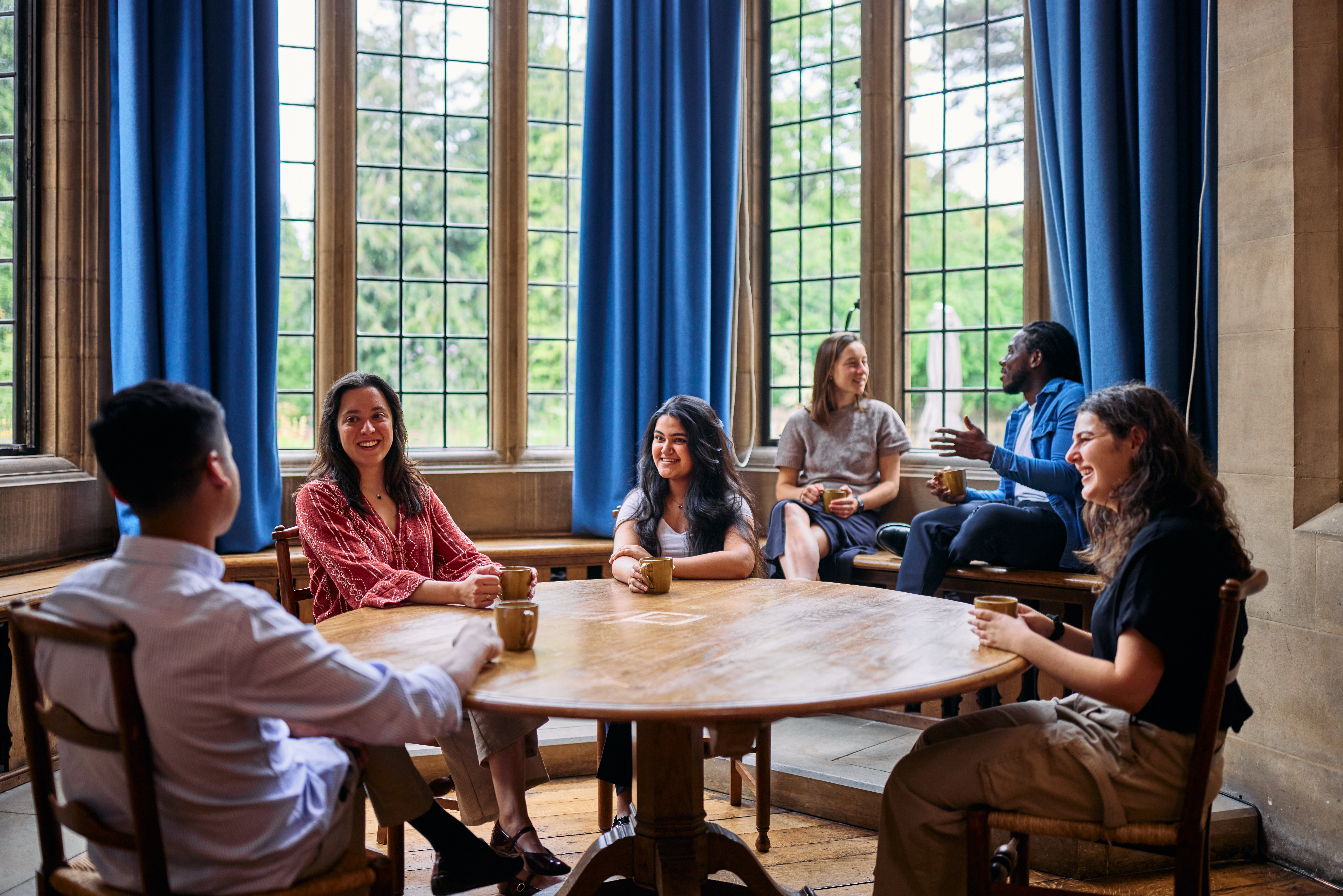A group of Rhodes Scholars engaged in lively discussion around a table in an Oxford library, with wooden panelling and blue curtains.