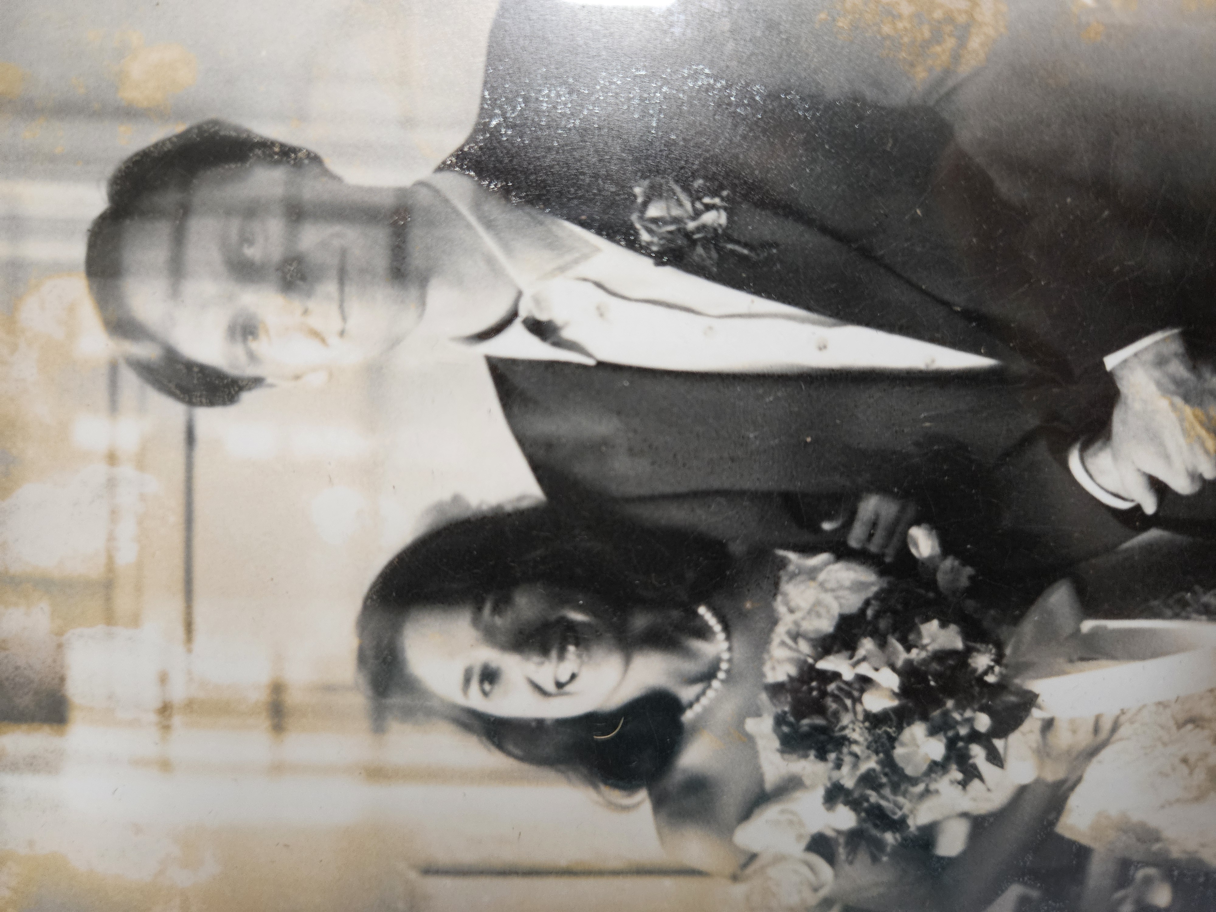 Black and white photo of a married couple. To the left is a lady smiling wearing a white dress, pearl necklace and holding a bouquet. To the right is a young man in a shirt and blazer and rose in the pocket.