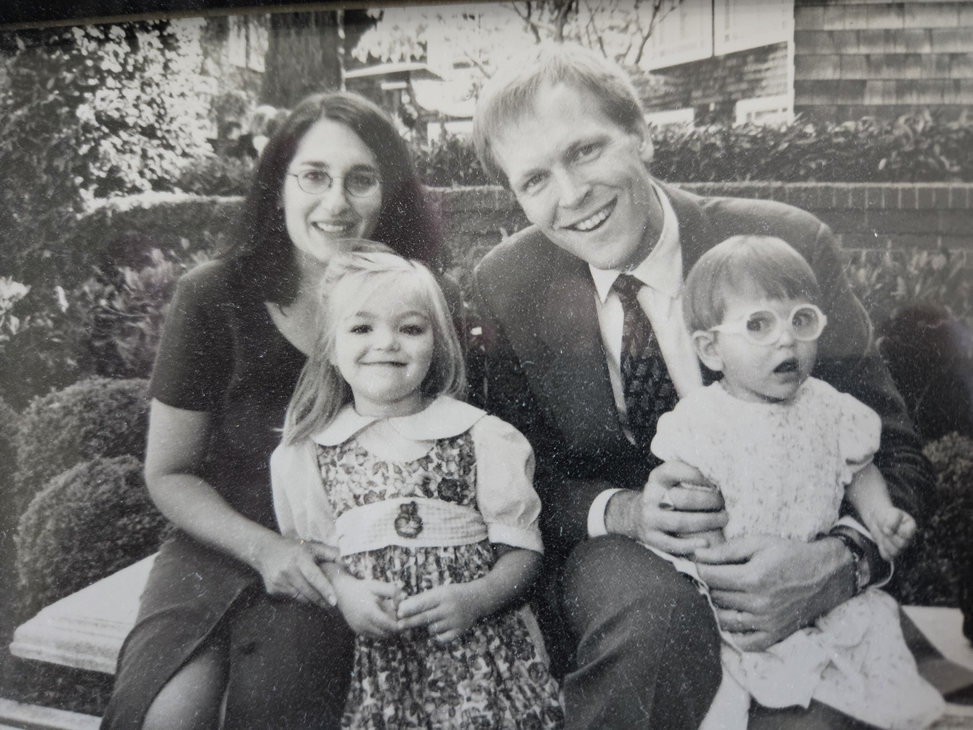 Black and white family photo taken on a bench. A mother and a father sit and smile on the bench holding their younger children, one girl and one boy.