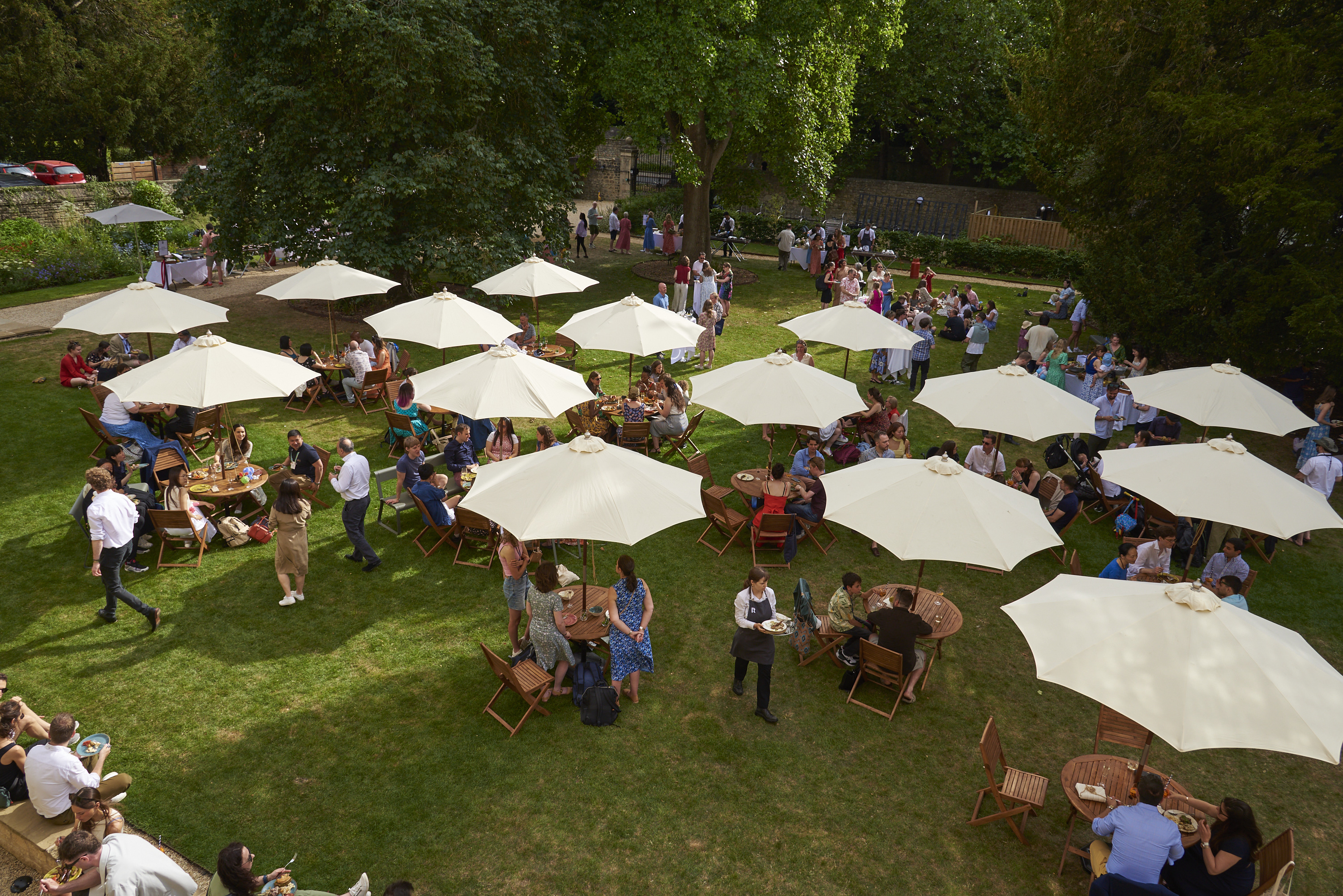 Garden party guests on the Rhodes House West Lawn with outdoor furniture and umbrellas, as seen from the first floor. Photo courtesy of Nasir Hamid.