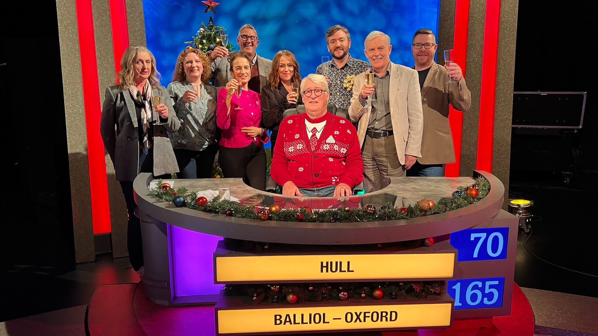 Elizabeth stood with a group of friends in front of a University Challenge podium reading 'Hull' and 'Balliol-Oxford'. There are festive decorations around them, with the host wearing a Christmas jumper and a Christmas tree displayed behind the contestants. Everyone is holding a glass of prosecco and is holding it up to the camera, smiling.