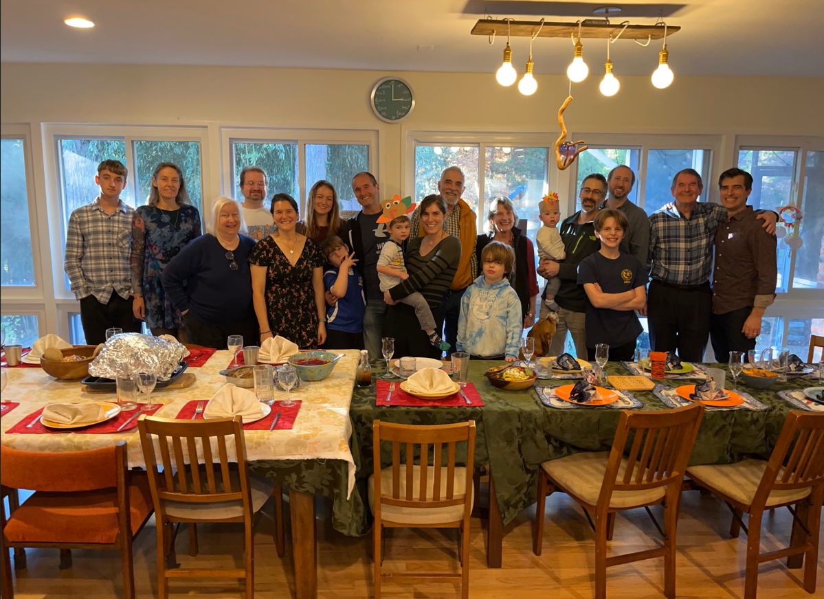 The picture shows a larger family standing behind a table. There are plates, cutlery and food on the table.