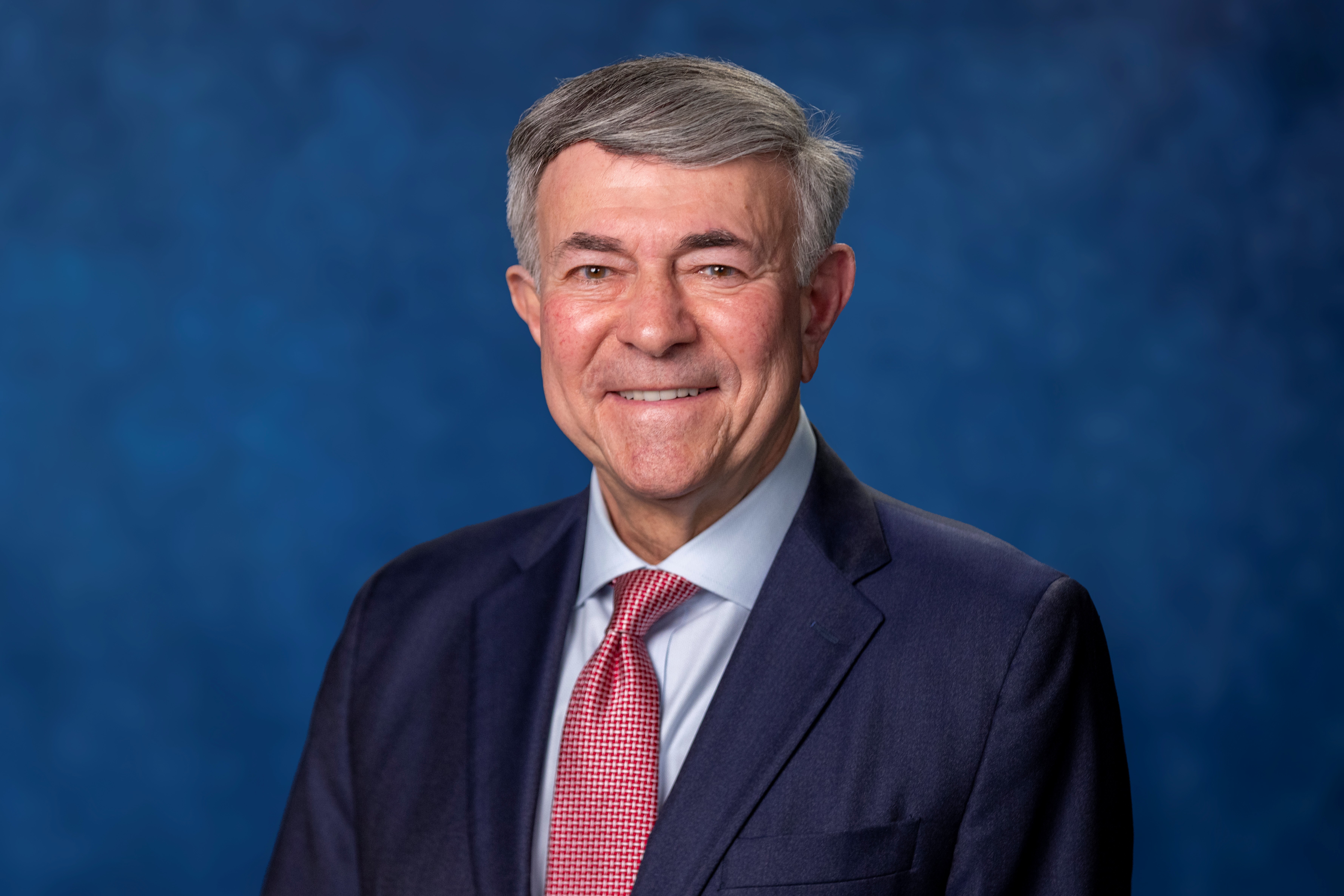 Bob wearing a black suit, smiling into camera, in front of a blue background.