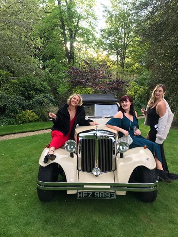 Three women are sitting on an old white car wearing formal dress