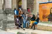 A group of seven Rhodes Scholars talking outside the steps of Rhodes House, in front of the colourful hoardings. 
