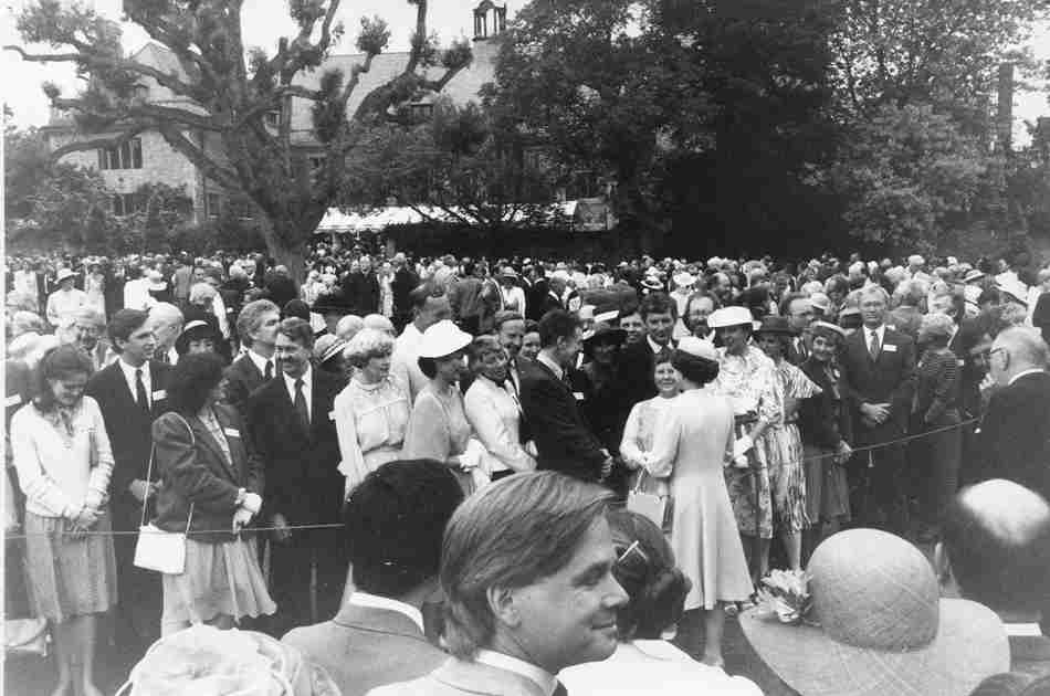 A black and white photograph showing a crowd of smartly dressed people waiting to greet the Queen. Many of the women are wearing hats. The Queen herself wears a long pale dress coat and pillbox hat.