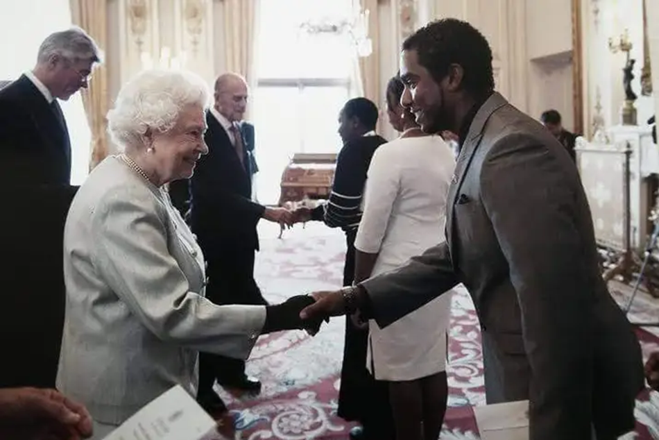 Kiron Neale shakes hands with the Queen inside Buckingham Palace. She wears a light grey suit and pearls, he wears a dark grey suit. They both smile and make direct eye contact. Prince Philip is shown shaking hands with another guest in the background.
