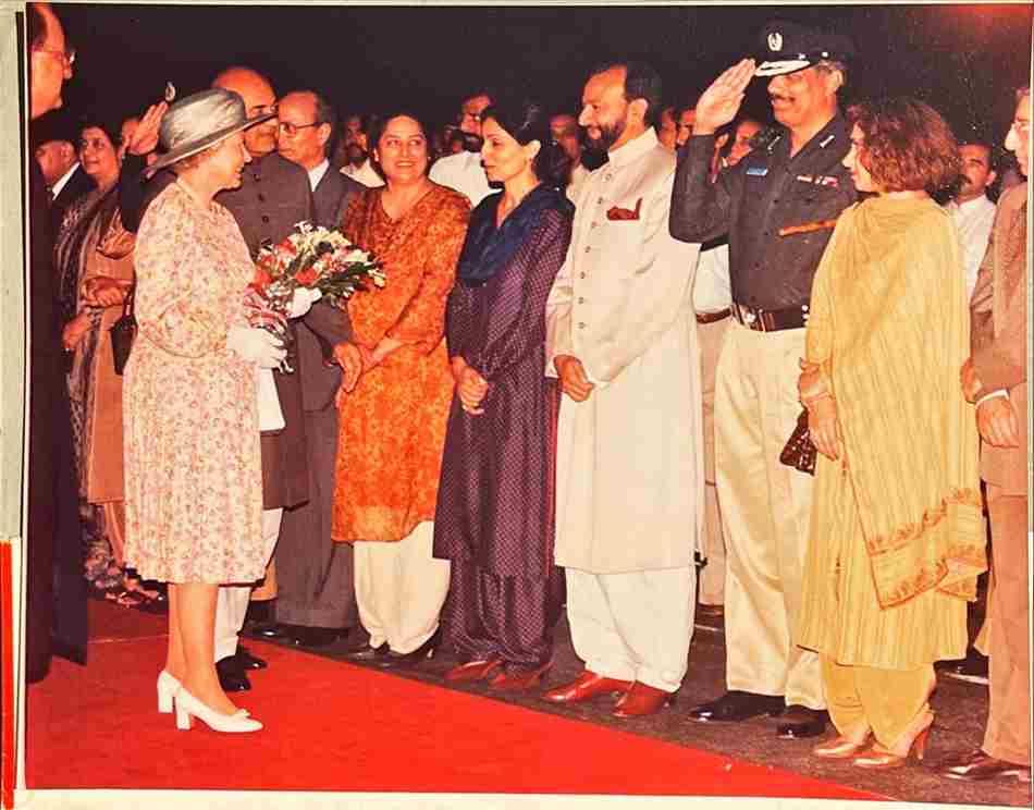 A group of people welcome the Queen to Lahore Airport. The Queen holds a bunch of flowers and wears a silver hat, floral dress and white shoes and gloves. She smiles at Dr Samia Malik, wearing purple, and Athar Tahir, wearing cream.
