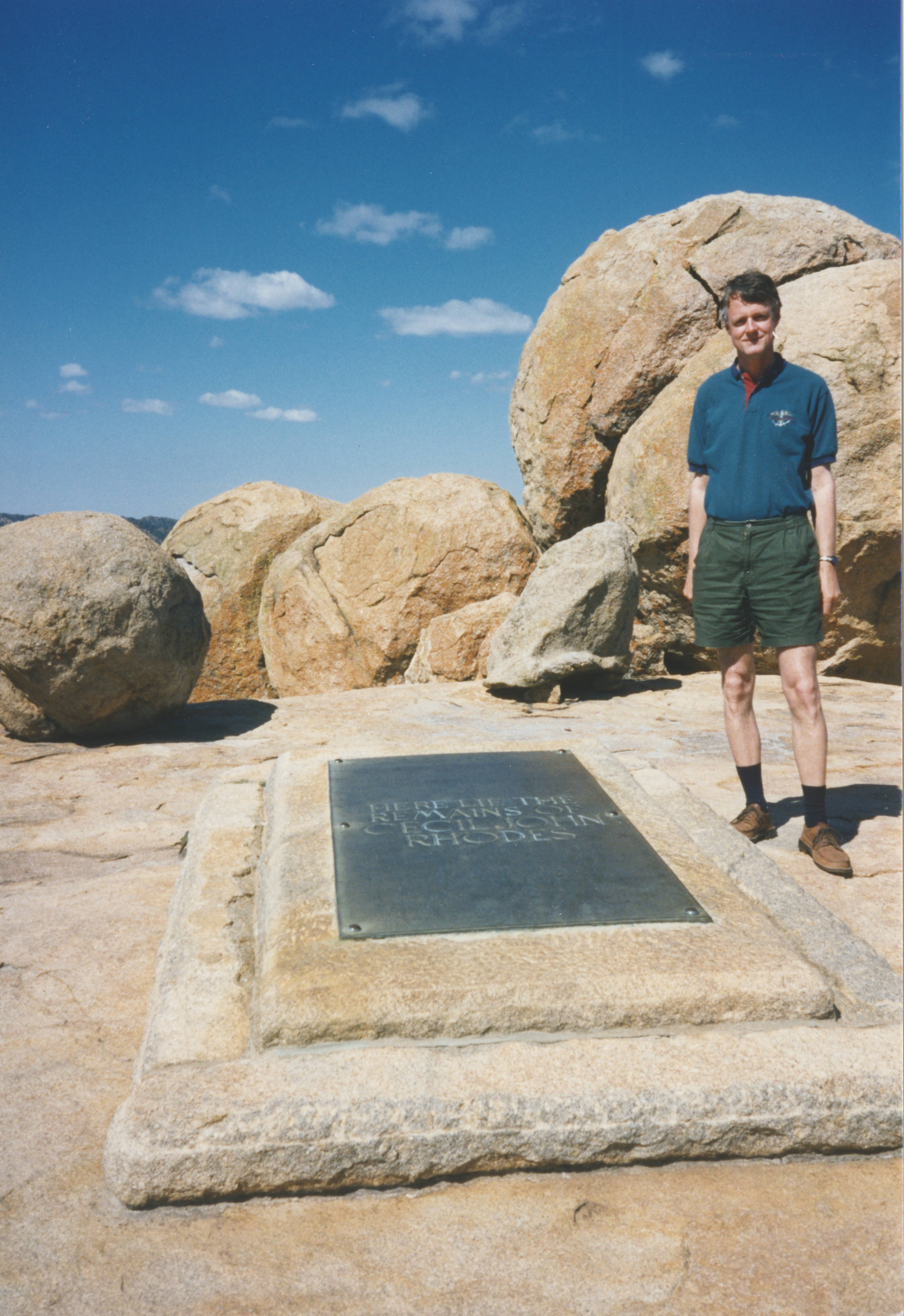 Jürgen is stood to the right of a grave, and at the top of a hill. Behind him are large rocks and a blue, slightly cloudy sky.