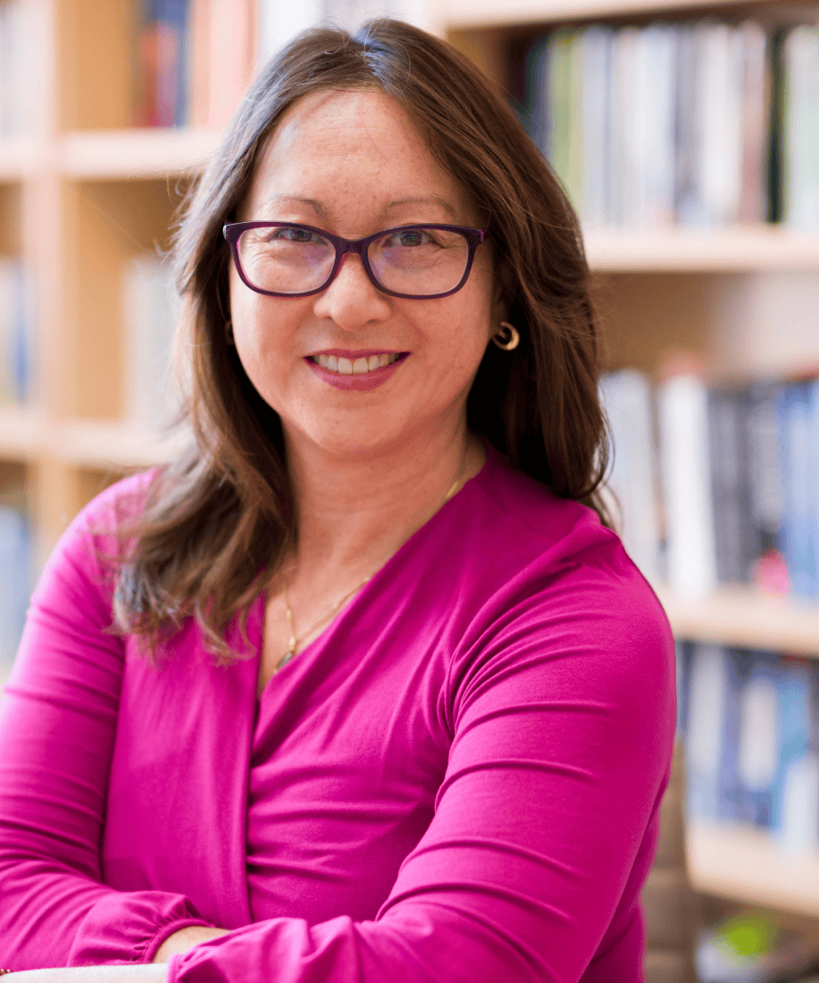 Portrait photograph of Karen. She wears a bright pink top, is posing in glasses, smiling. Behind her is a wooden bookshelf filled with books.