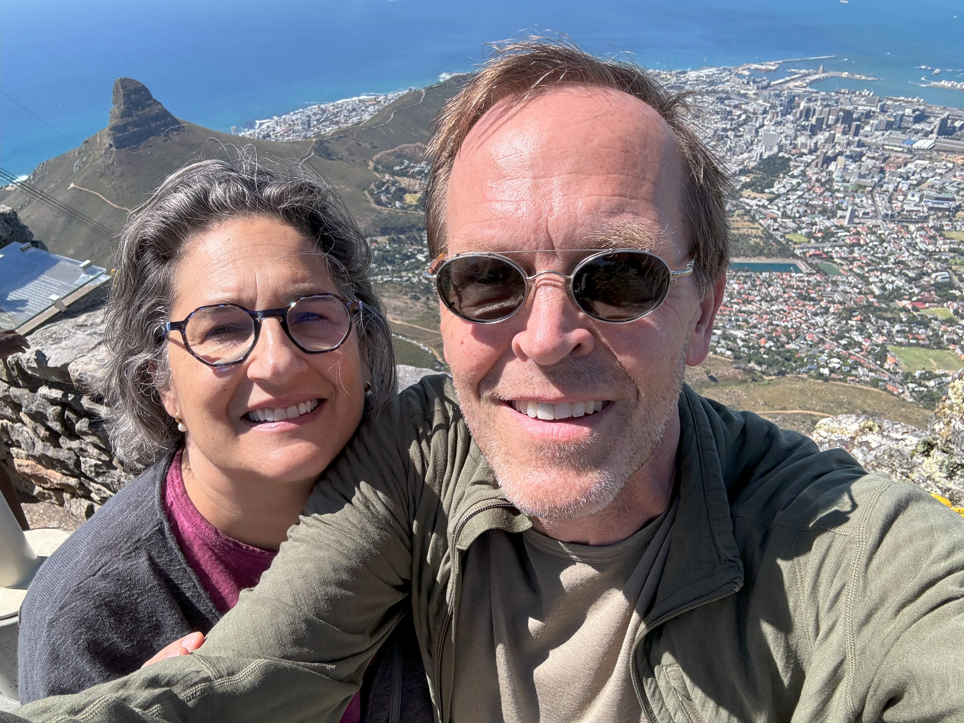 Selfie photo of Eric and his wife. They appear on the top of a mountain as behind them we see a city view, hills, and the ocean. They are both smiling.
