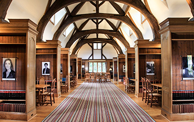 An image of the Rhodes Roseberry Room, which is a long, wooden structure with a vaulted ceiling, a striped carpet down the middle, and wooden sections on both sides with tables and chairs for study. 