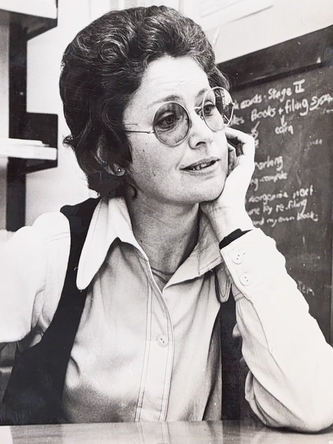 Black and white photograph of Elizabeth. She is leaning with her elbow on a table, and is holding her face. She is wearing large round glassses, and a blackboard can bes een behind her.