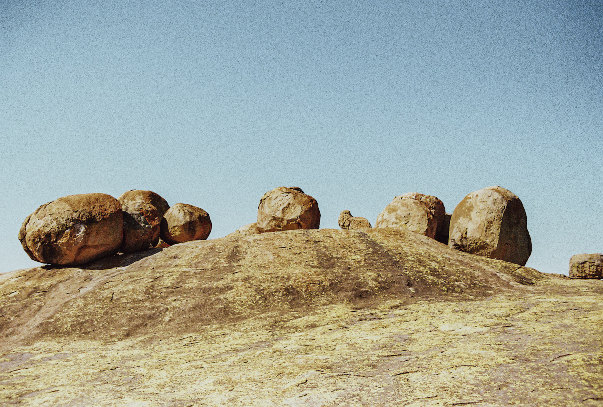 Eight granite boulders rest along a broad lipped granite rise in broad blue-skyed daylight at high-noon casting short shadows on the lichenified rock below