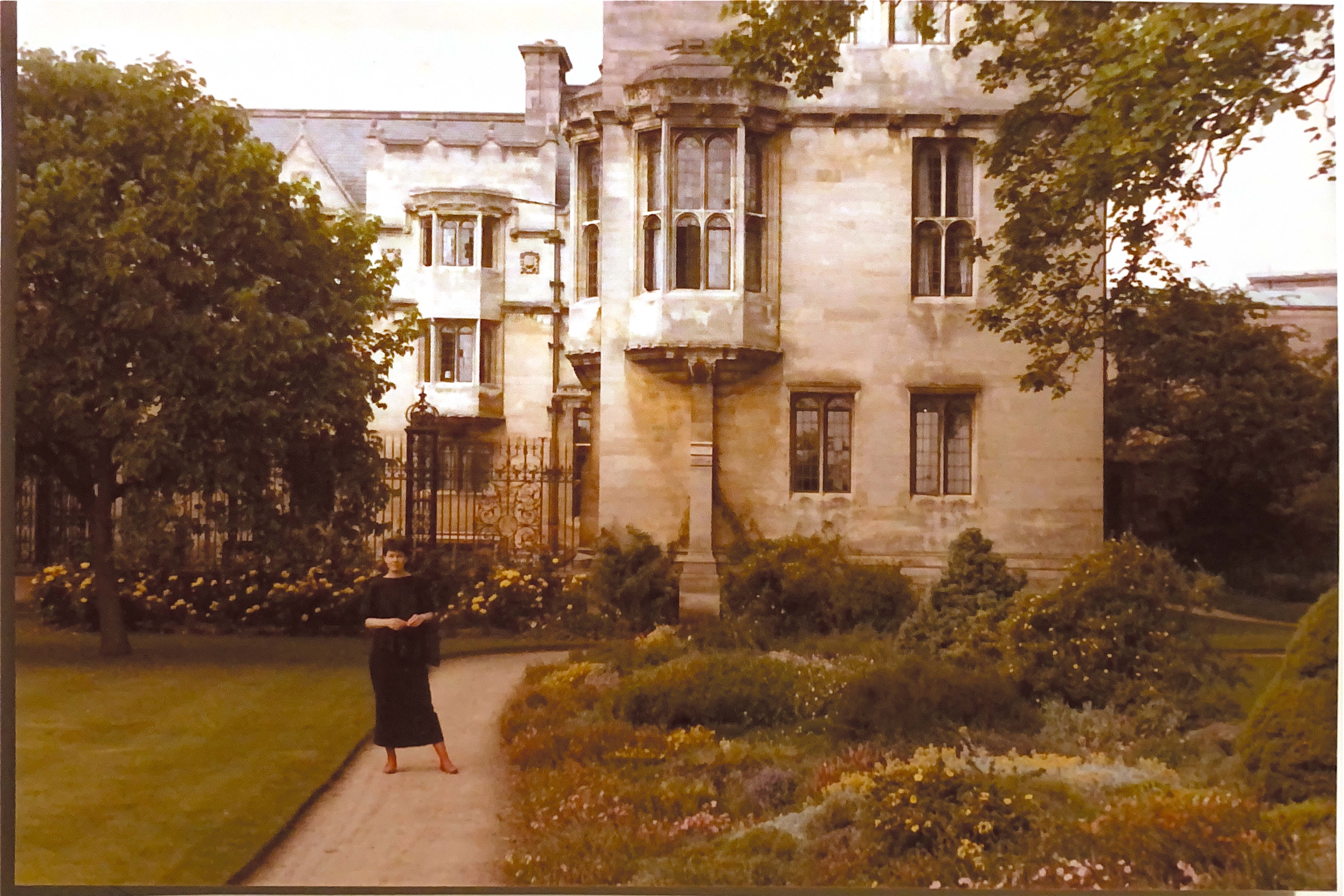 Susan is stood in front of a large college building, with the gardens in bloom around her.