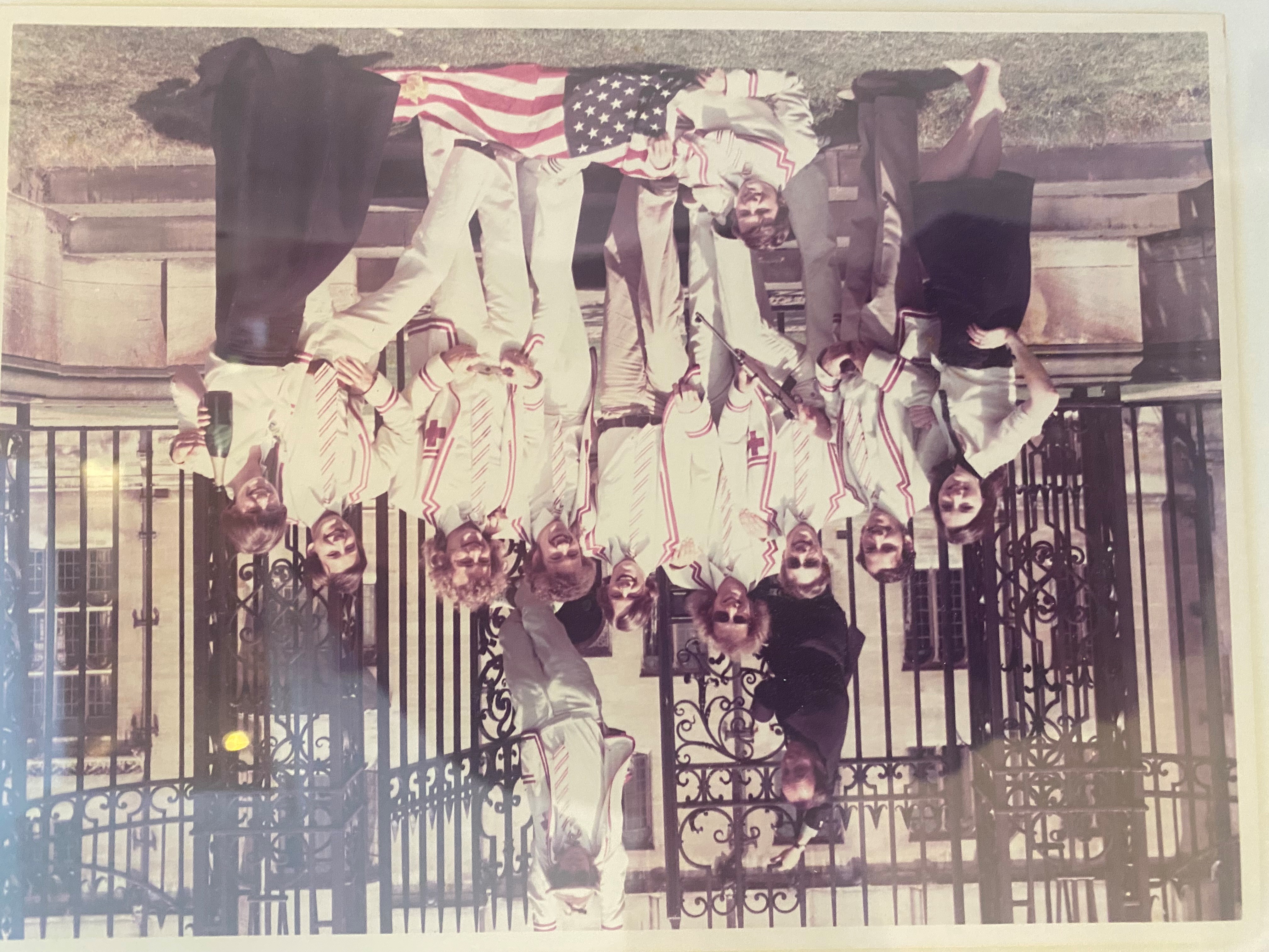 Sepia photo of sports team all stood smiling before a gate, with Nick leaning on the floor holding a USA flag.