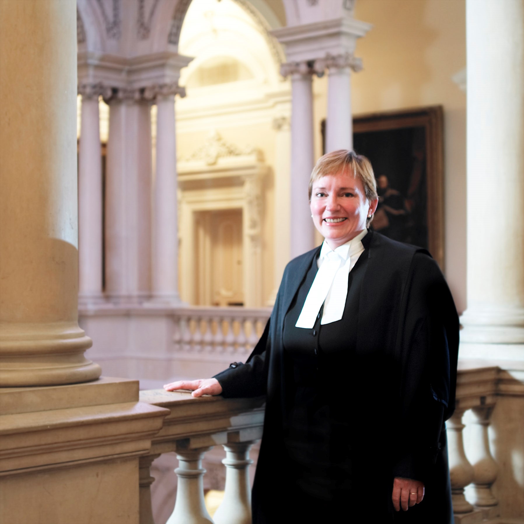 Eileen stood next to a bannister, wearing her black and white judges outift. She is smiling into the camera, and there is a grand hall with columns and a painting (unclear) behind her.