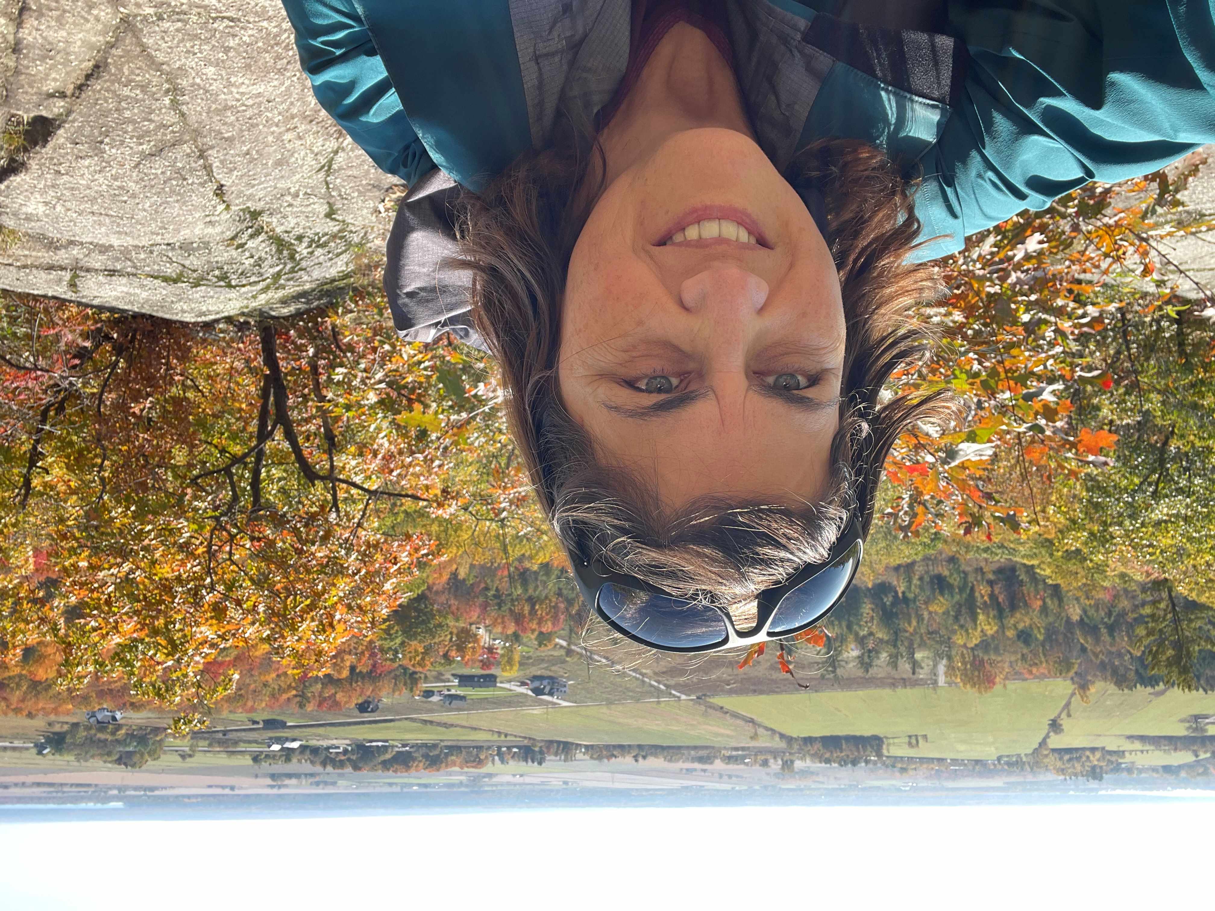 Carol taking a selfie on a hill, smiling, with a field behind her.