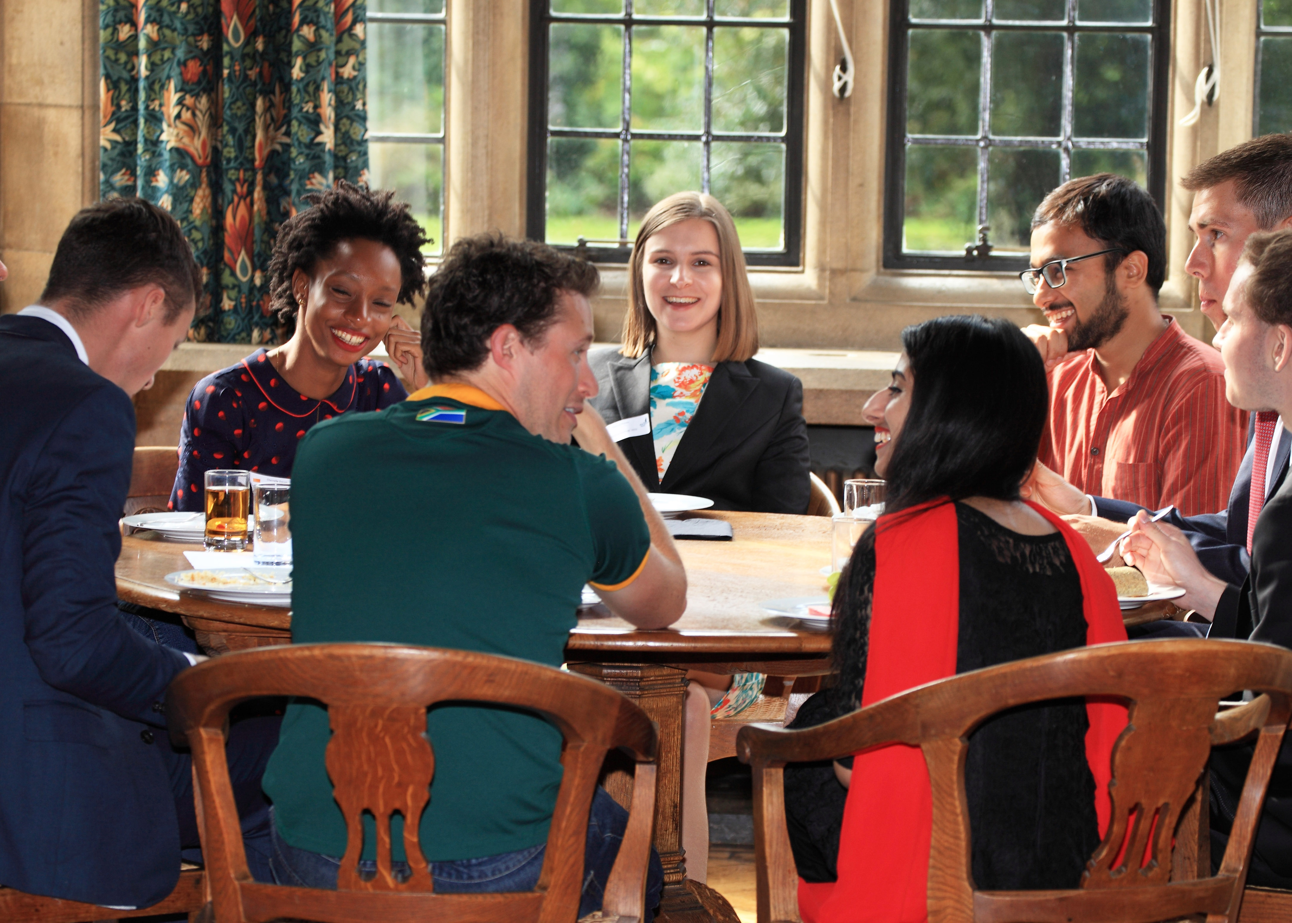 A group of eight scholars are gathered around a wooden table in a circle smiling and talking to each other. They are indoors next to several windows in a stone wall. 