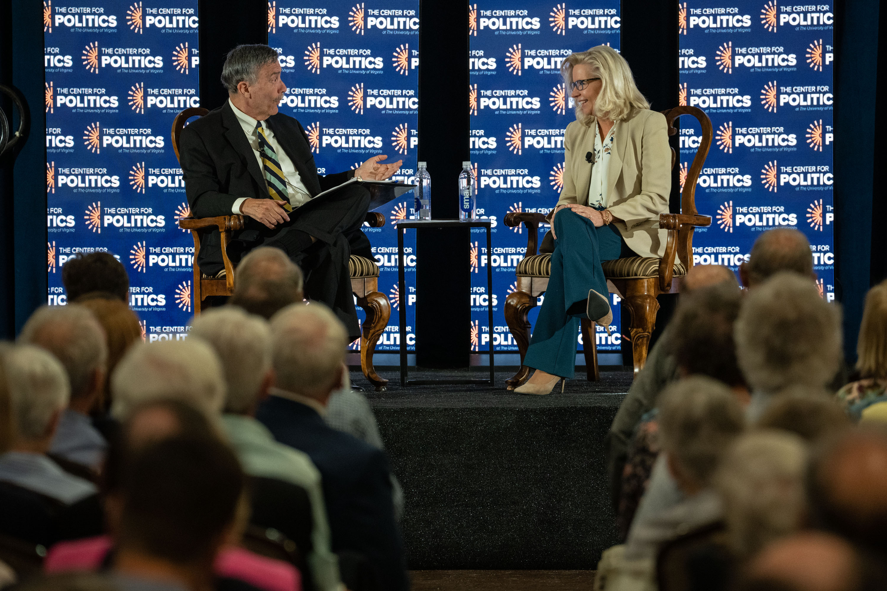 Sabato and Liz are sat on chairs facing each other with a crowd before them. They are mid conversation, and the banner in the background reads 'The Center for Politics'.