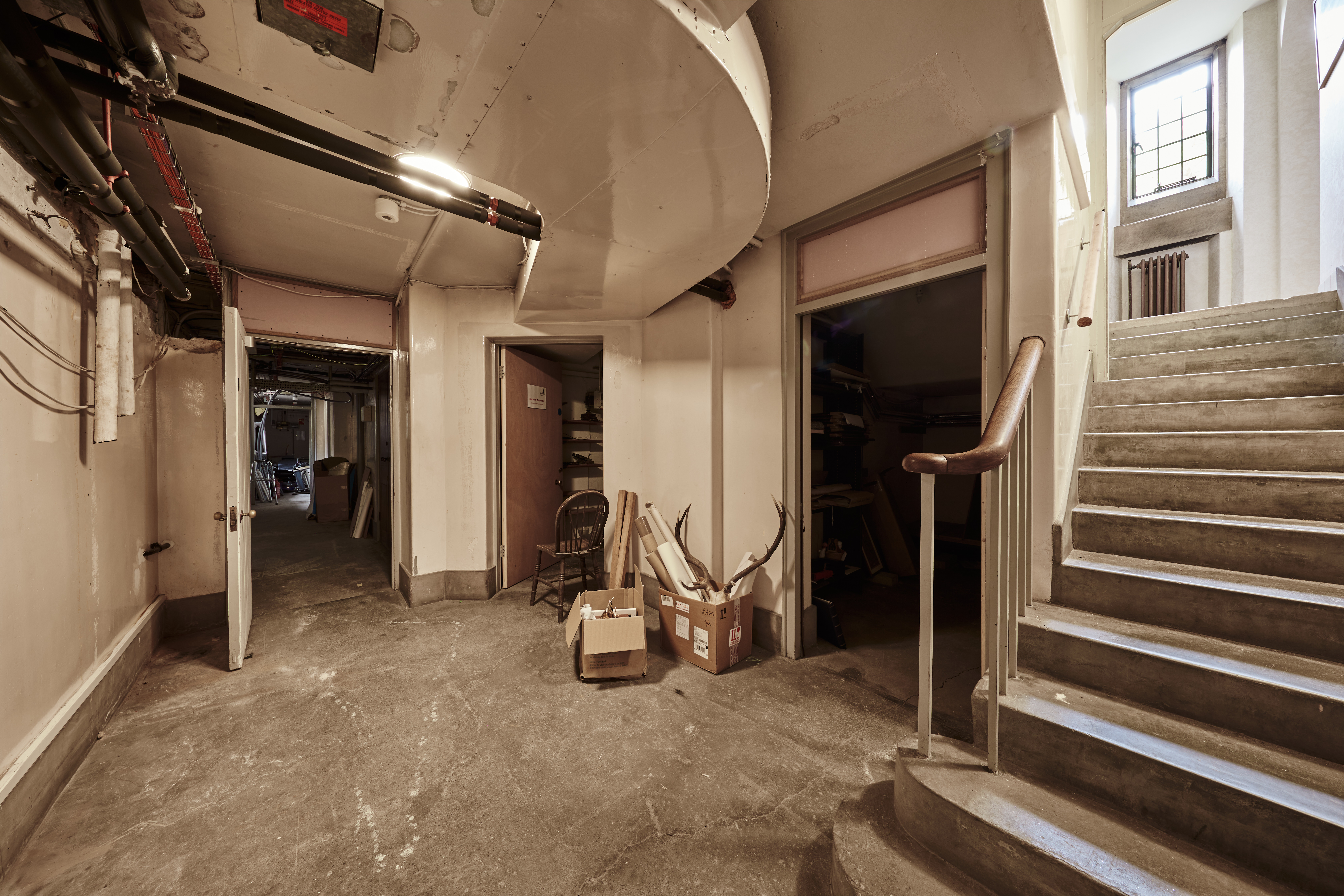 Image of the Rhodes House Basement. There is a stairwell on the right leading to the basement, which is empty and shows a couple of storage boxes in the back.