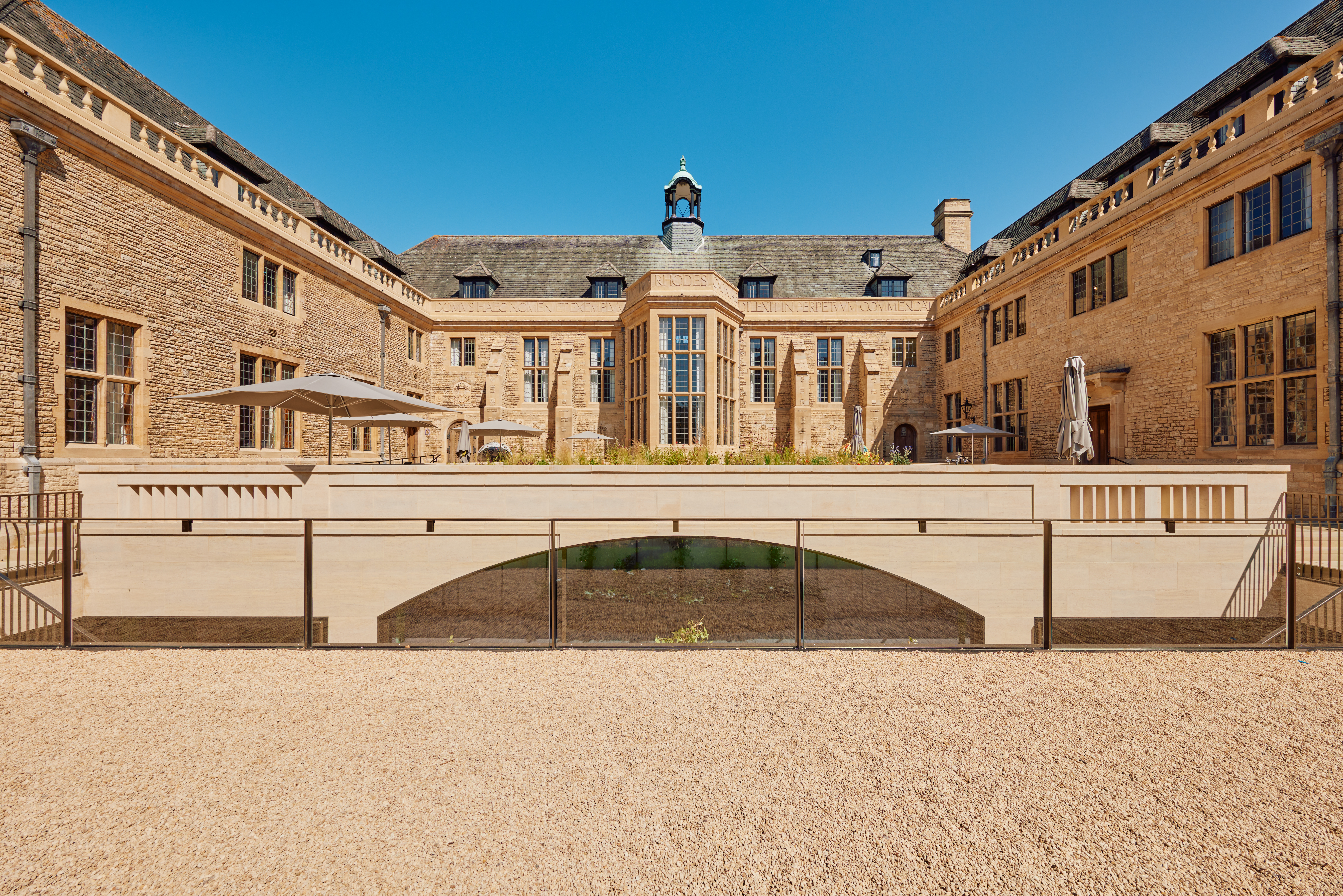 An image of the Rhodes House convening centre exterior. It is outdoors, surrounded by the stone walls of the building, and there are parasols in the seating area. It is sunny and there is a clear blue sky. 
