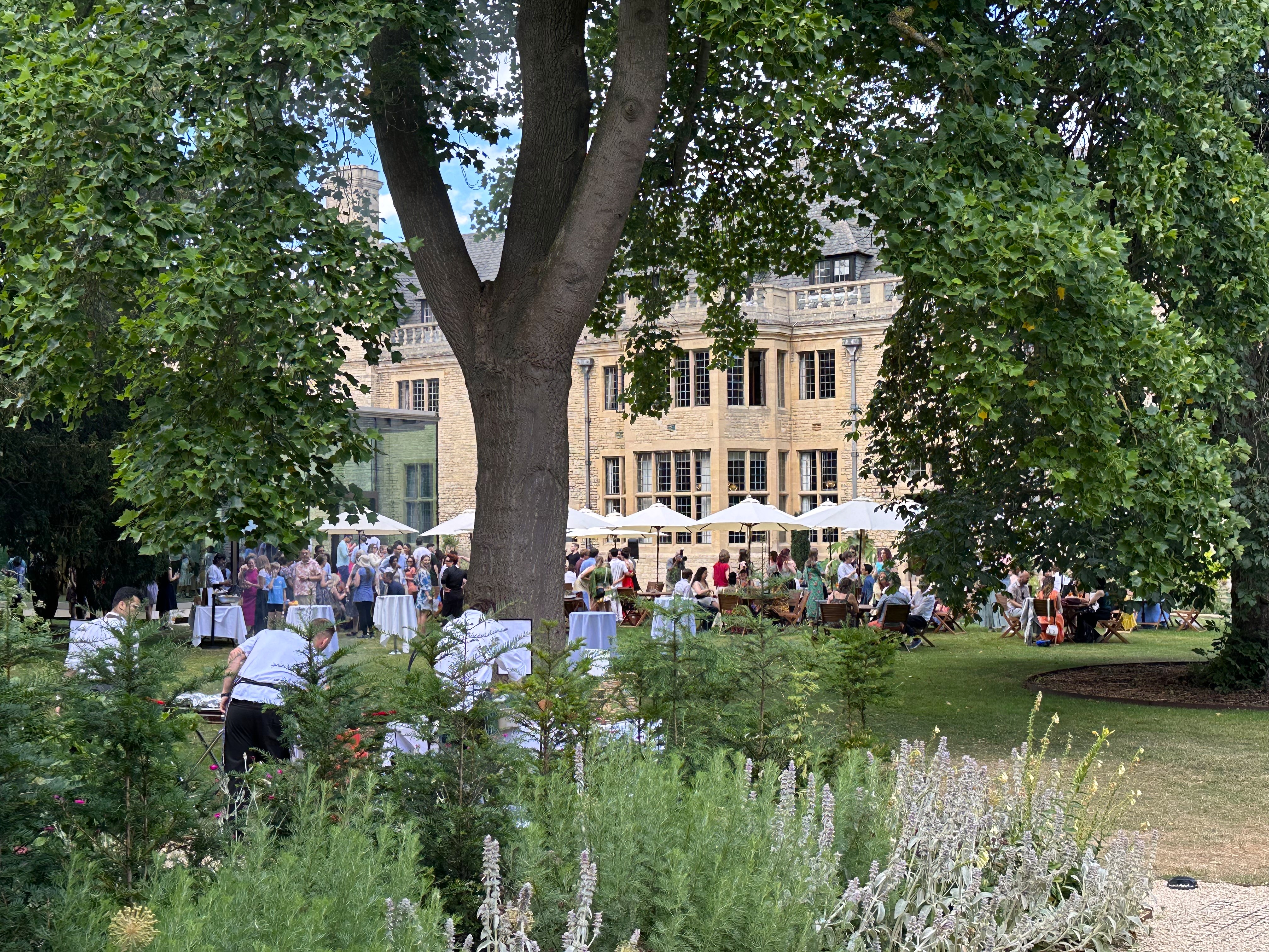Guests at a Rhodes House BBQ with the chefs preparing food under the trees.