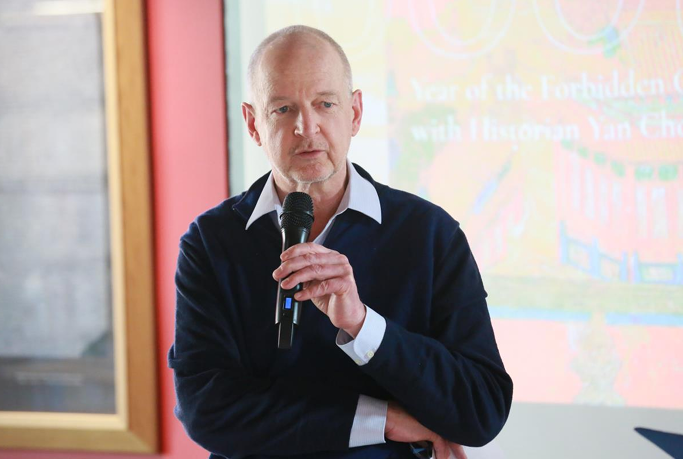 Malcolm is pictured holding a camera and appears to be speaking to an audience. The background display behind him is not fully eligible, but the words 'Year of the Forbidden' and 'with Historian Yan' can be seen.