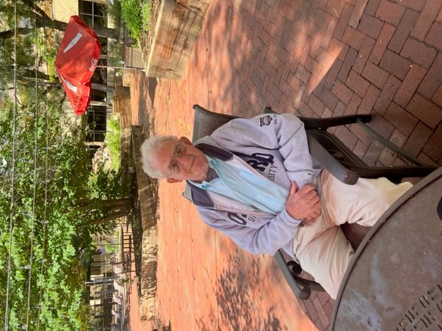 Joe sitting in a chair. He is wearing a sweatshirt over a sky-blue shirt and beige pants.