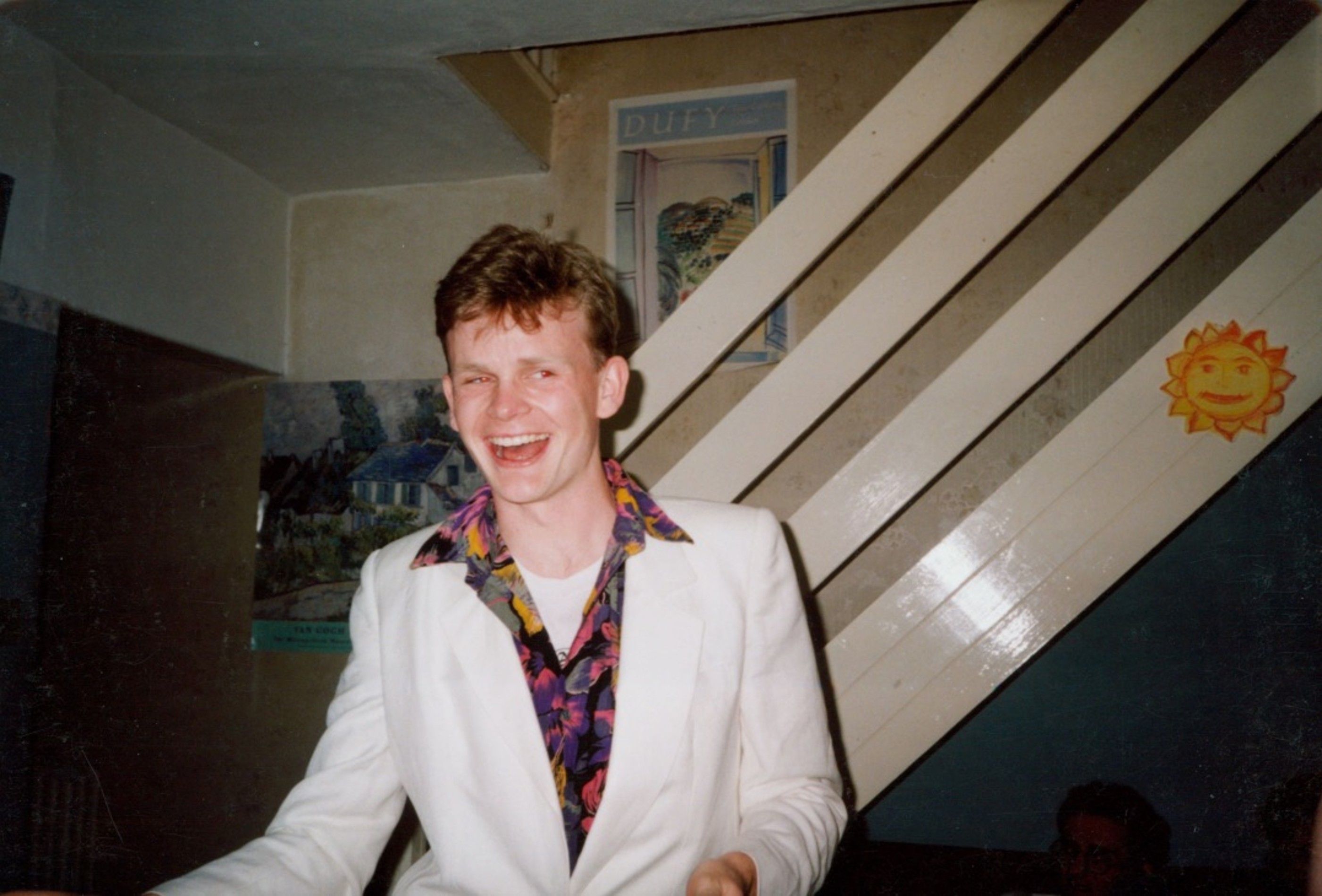 A picture showing Mark in a white jacket and a floral shirt. He is in a small room, with a staircase behind him, laughing at the camera. 