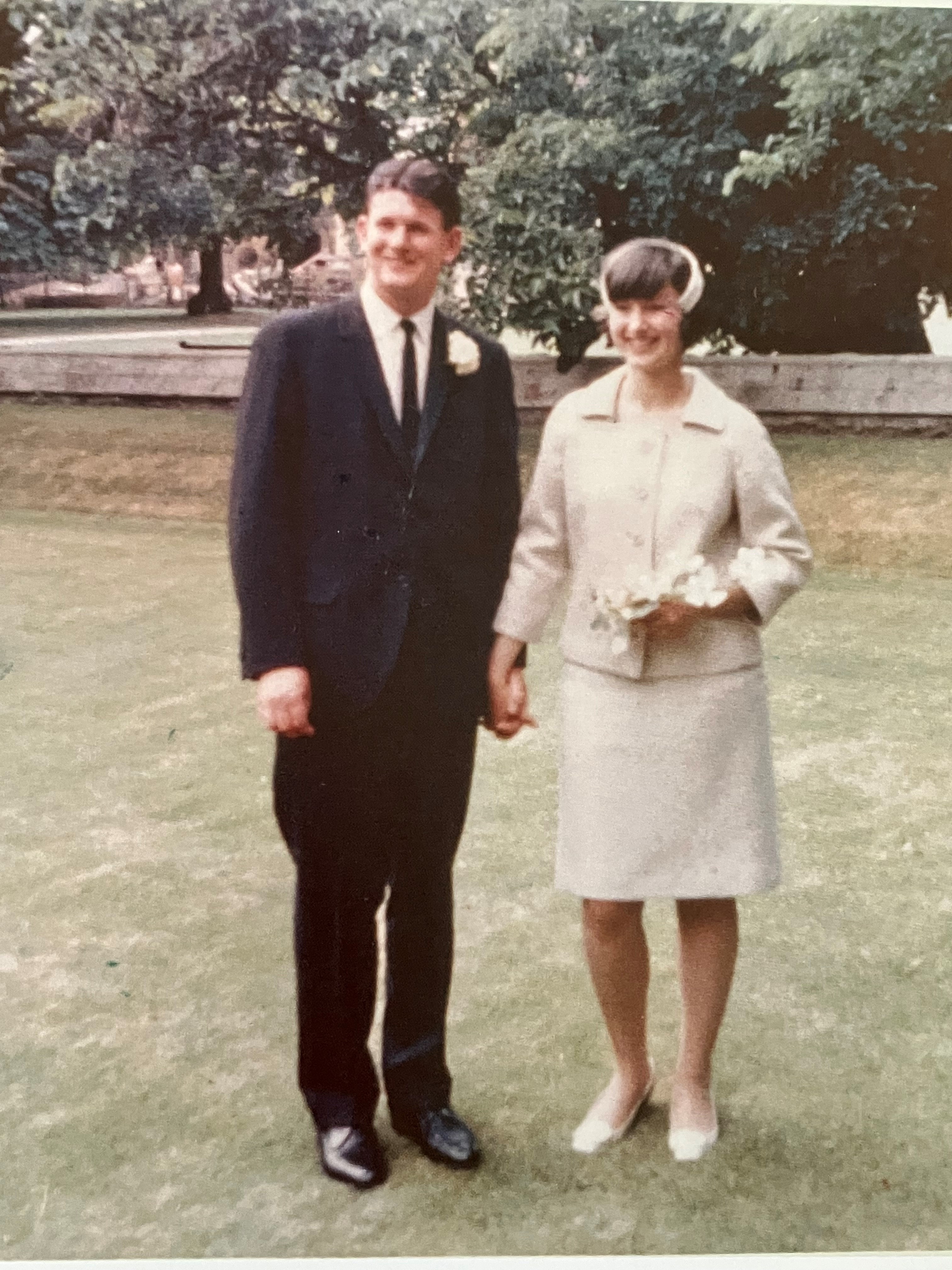 The picture shows Nick in formal attire with his wife in a white dress. They are standing on a lawn.