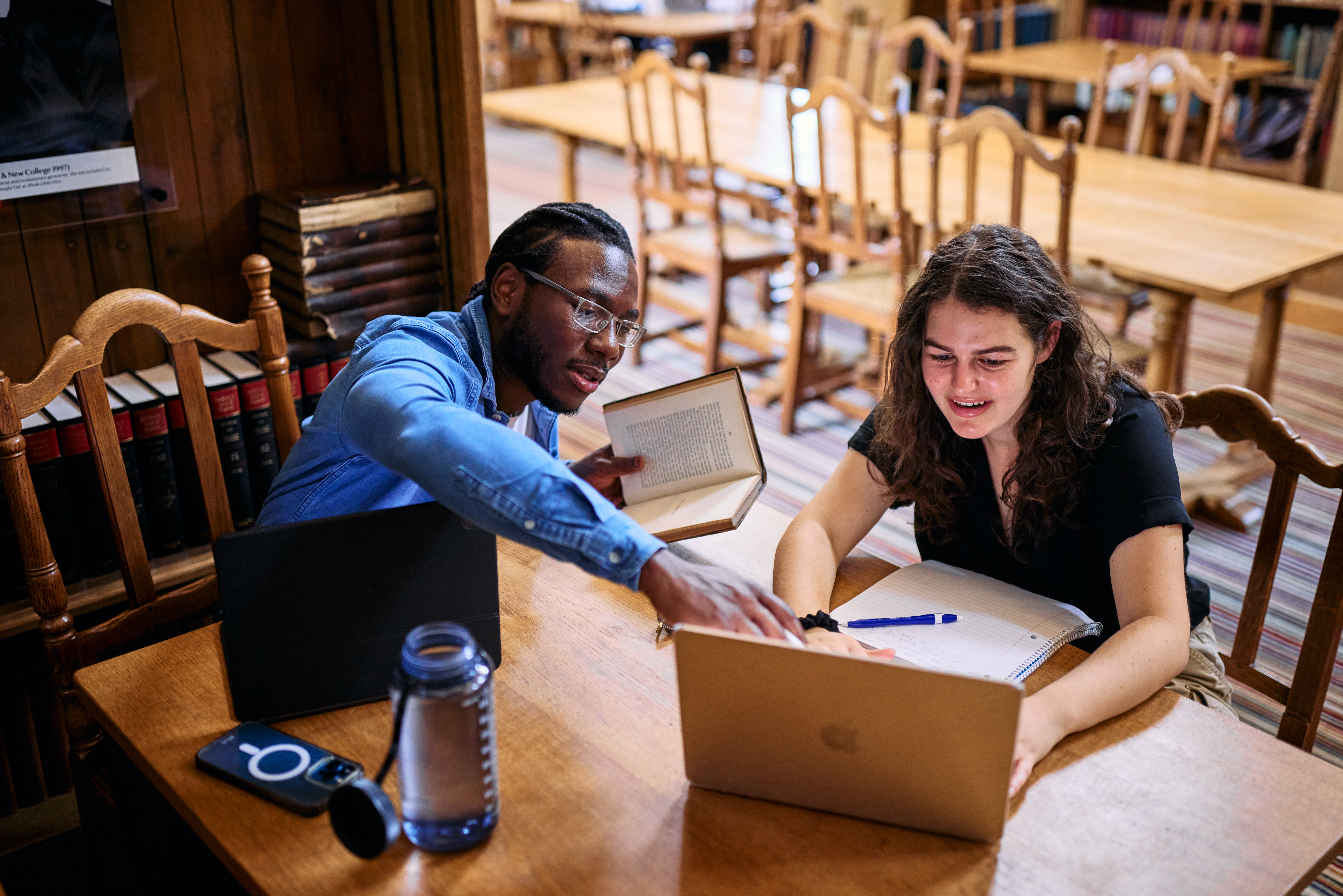 Two students working together in a library. One of them is holding open a book and pointing to the other's laptop screen, engaged in discussion.