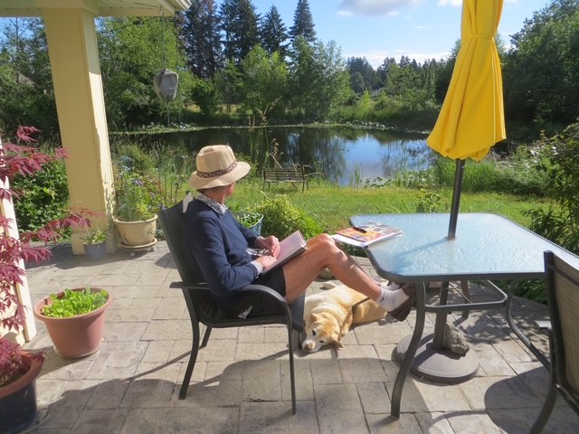 John sitting at a table holding a book next to a lake and a dog on the ground