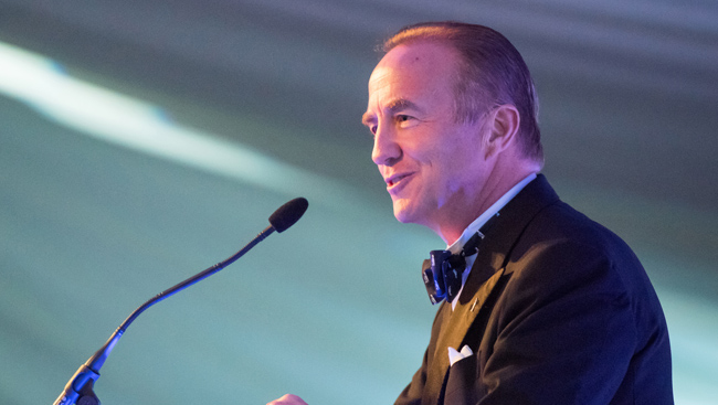 An image of John McCall MacBain, one of the Rhodes Trust's Second Century Founders. He is in a suit and bowtie speaking at a podium at an event.
