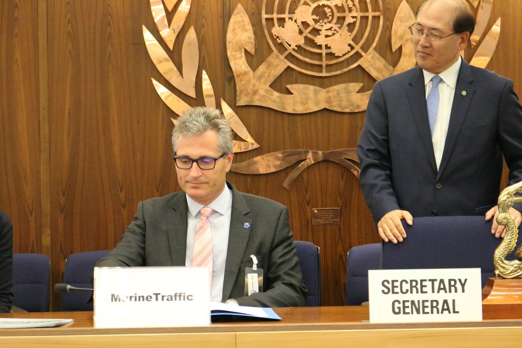 Argyris sat at a table. The sign before him reads 'MarineTraffic' and the gentleman standing to his right has a sign that reads 'Secretary General'