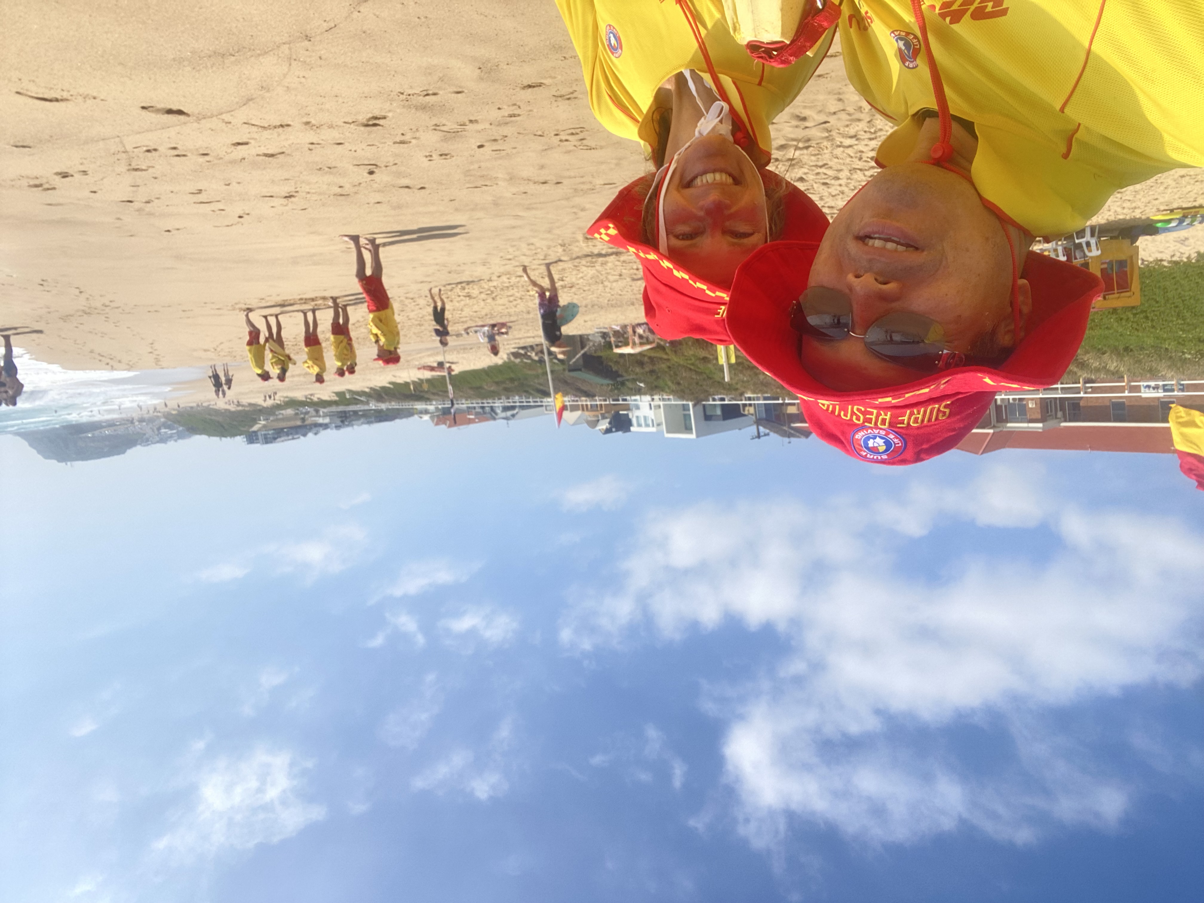 Selfie of Jenny and friend on a beach, blue sky and water, both wearing bright yellow and red outfits.