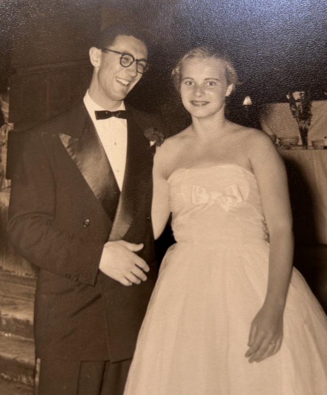 Black and white photo of Ken stood smiling in a tux to the left of a lady wearing a ballgown
