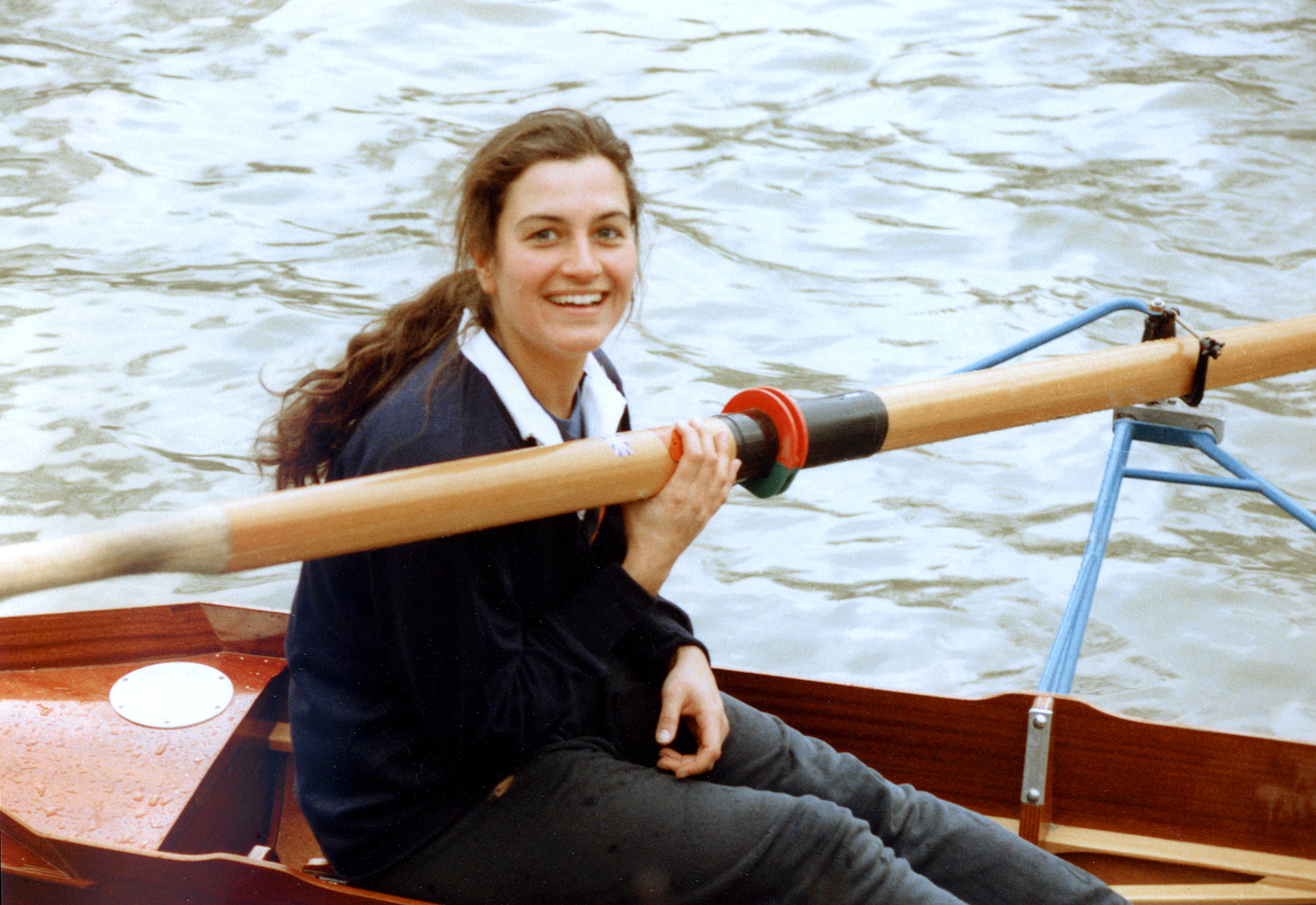Young Elizabeth sat in a rowing boat, hair in a ponytail, smiling, and holding an oar. The water is seen around her.