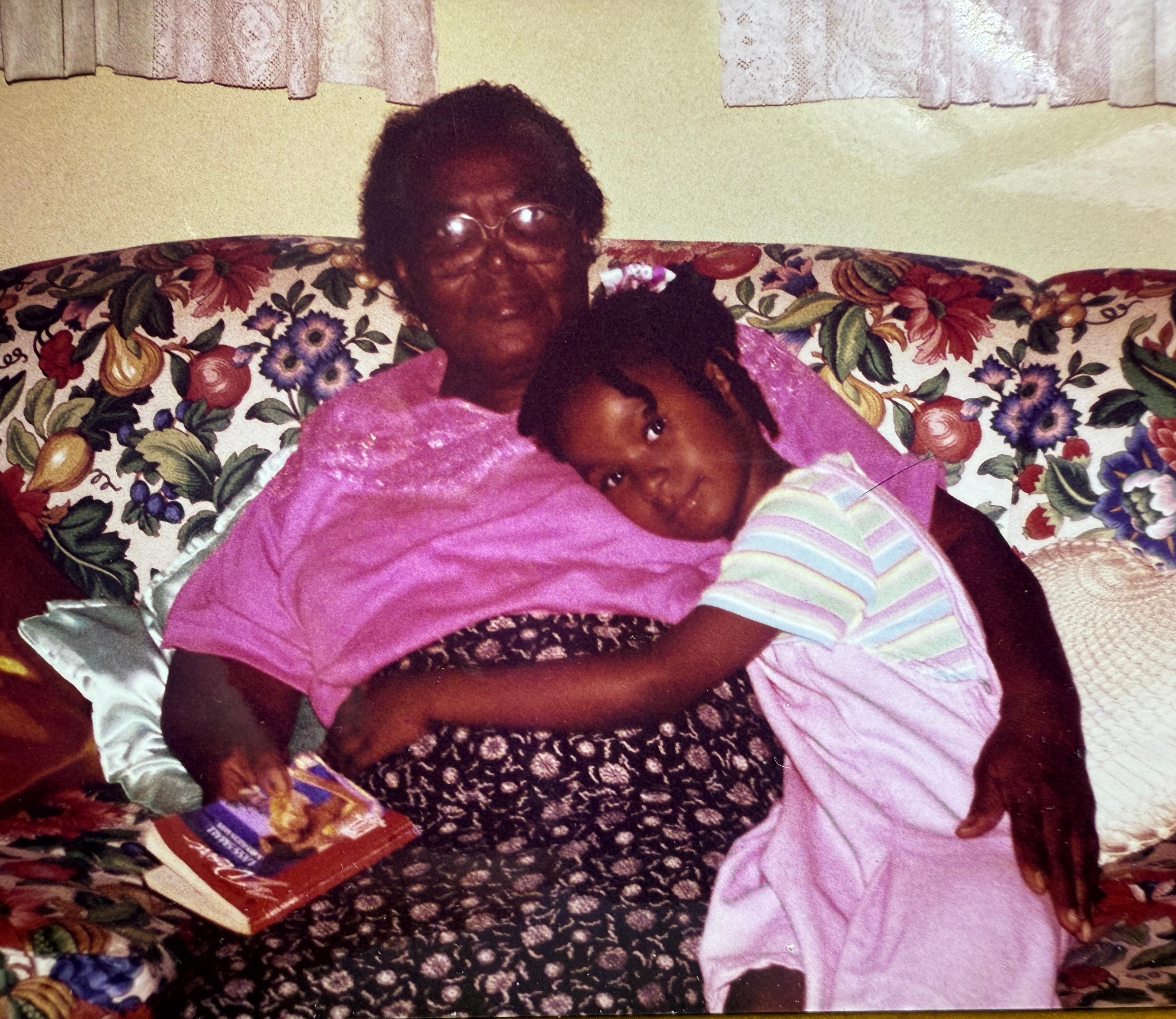 A baby Chloe holding onto an older woman who is holding a book and sitting on a sofa