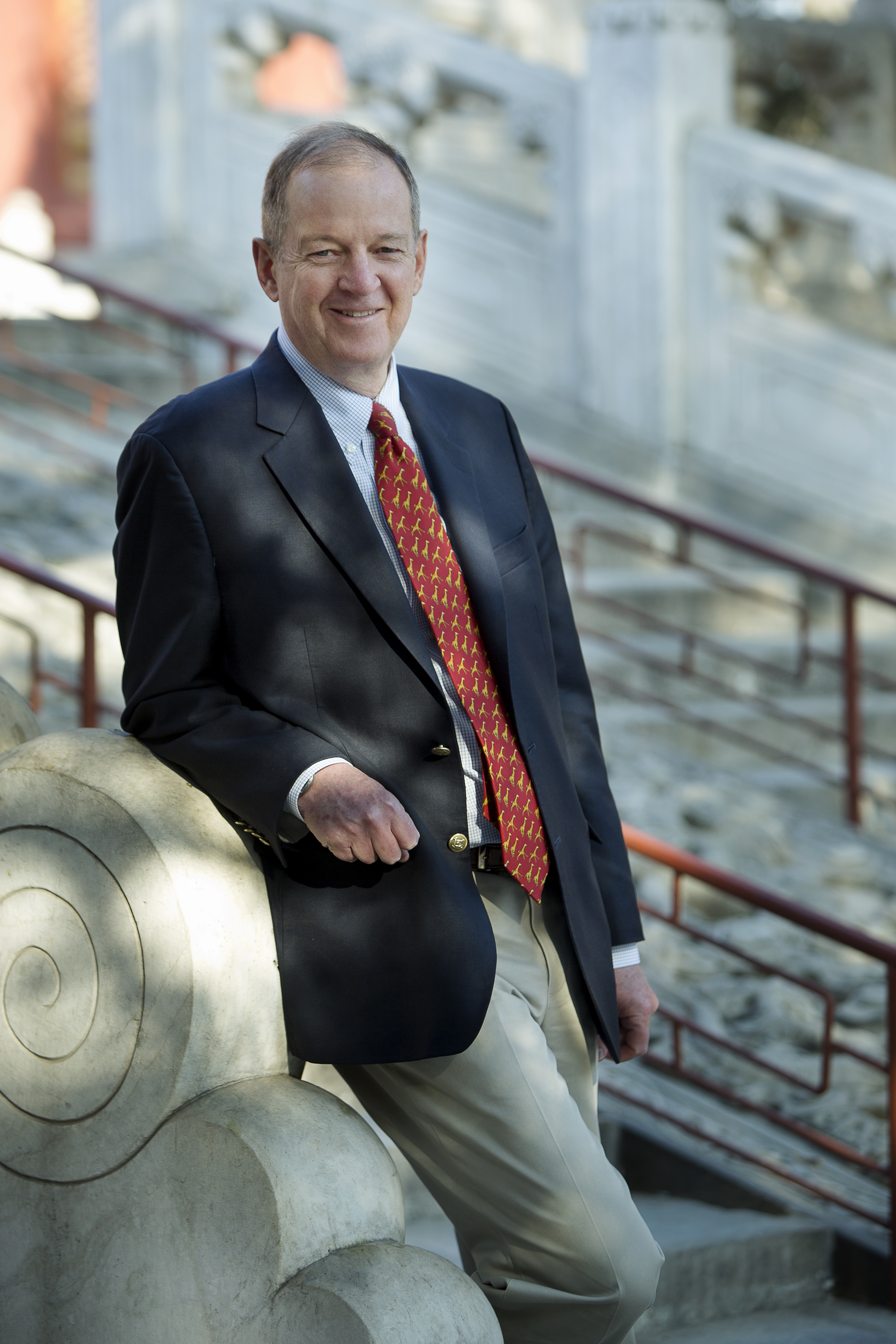 Portrait photo of Malcolm stood leaning against stairs, wearing a suit with a red tie. 