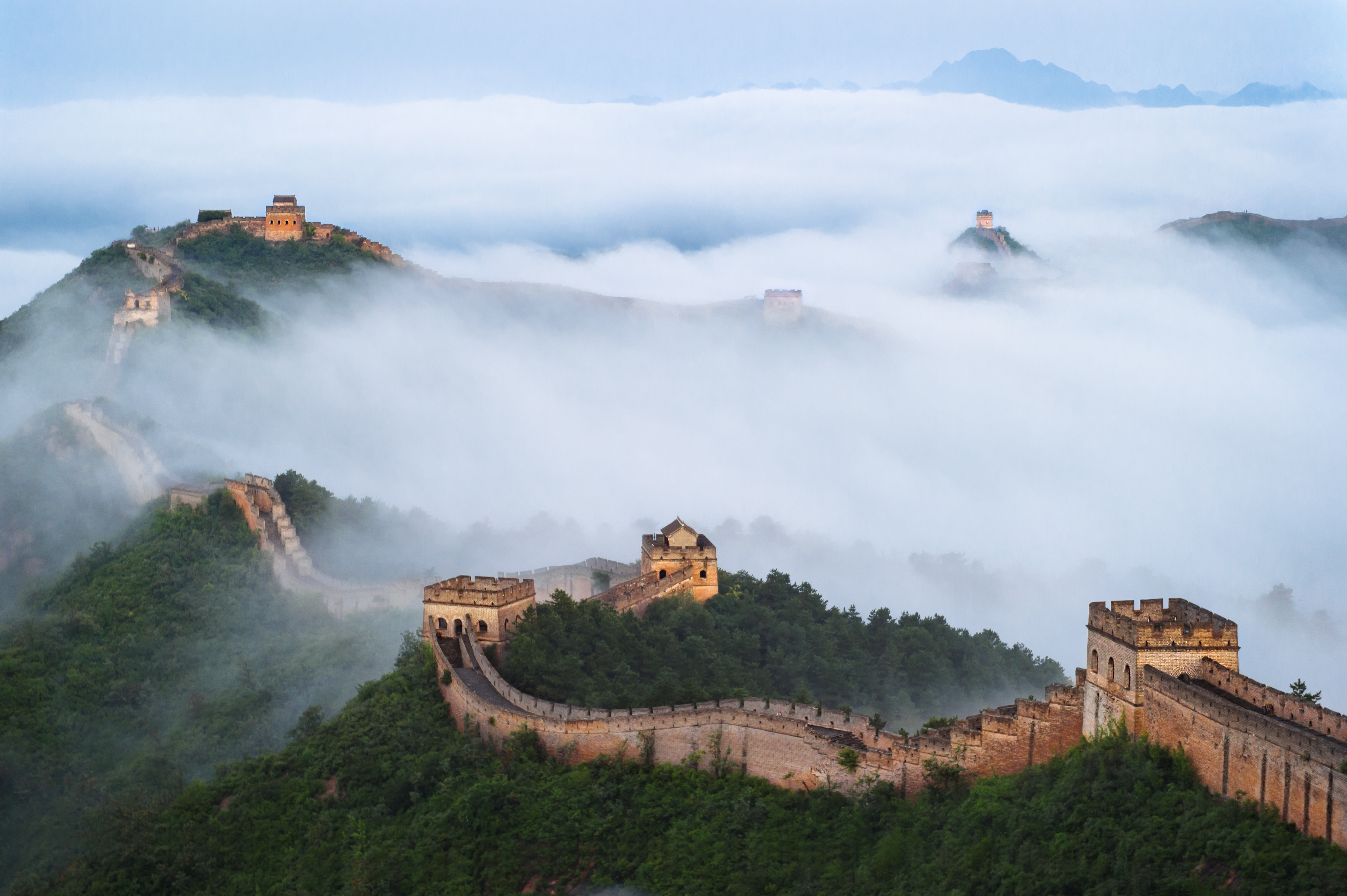 An image of the Great Wall of China from above. There are low clouds over the trees and structures.
