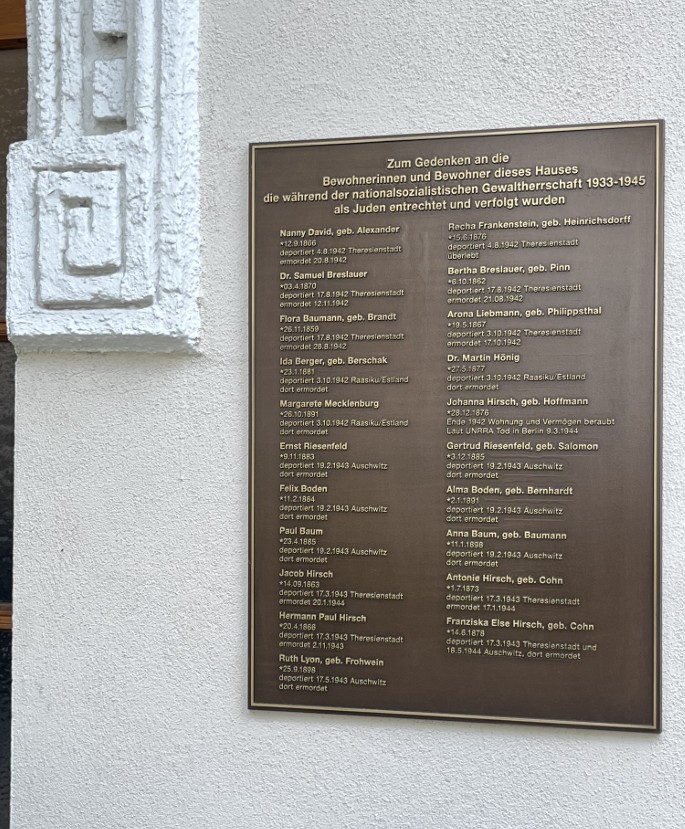 Brass Plate rememebering Jewish residents. It reads In German: 'Zum Gedenken an die Bewohnerinnen und Bewohner dieses Hauses die wahrend der nationsozialistischen Gewaltherrwschaft 1933-1945 als Juden entrechtet und verfolgt wurden. Underneath, 21 names are listed alongside their date of birth, date they were sent to a concentration camp, and date they died (if known).
