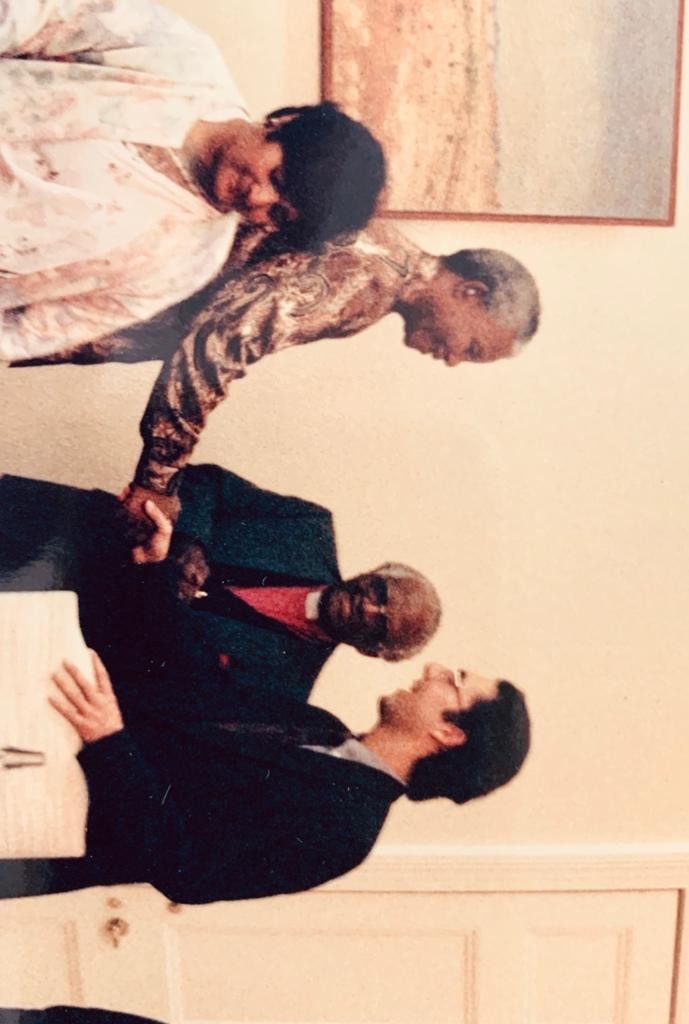 This image captures Paul stood to the right of Nelson Mandela and Archbishop Tutu. All in the image are smiling, and Paul shakes Nelson Mandela's hand whilst Archbishop Tutu is stood in the middle and smiling. A woman is walking past at the left of the image.