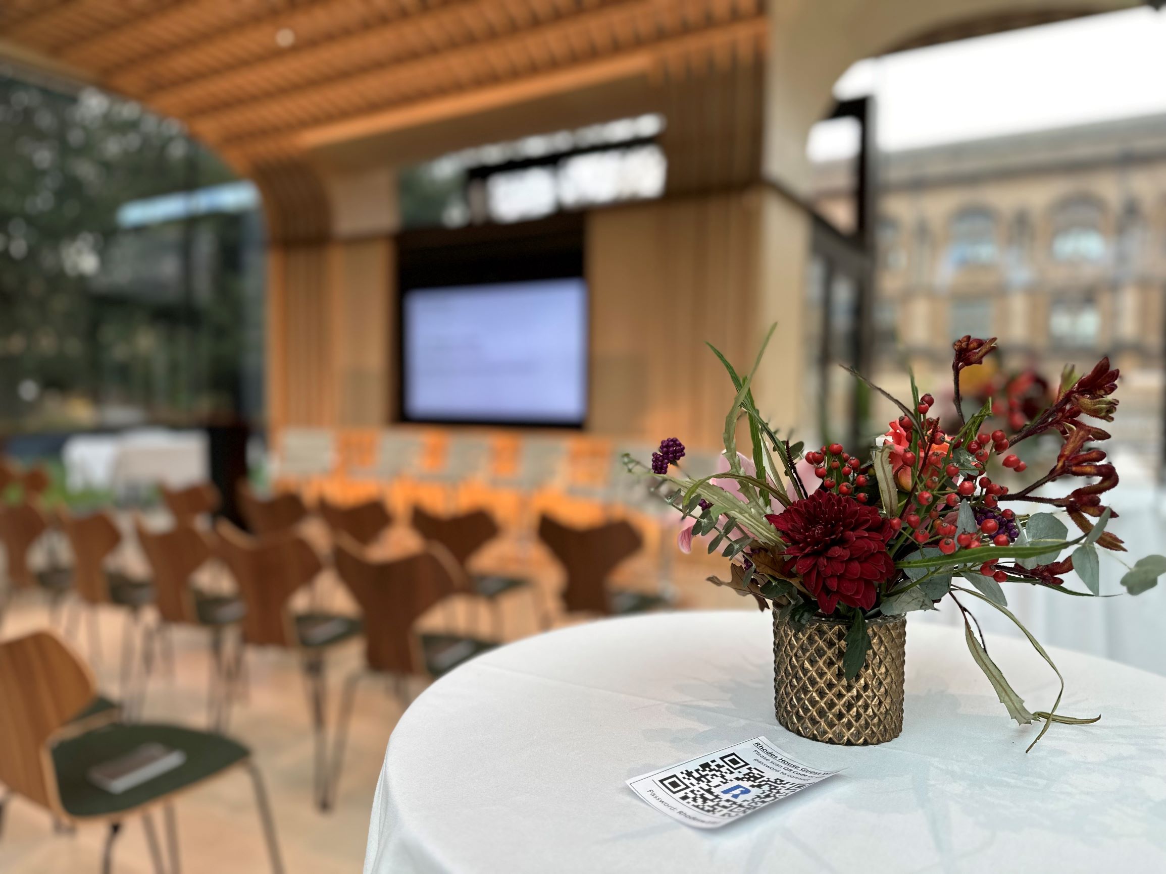 Rhodes House Glass Pavilion set up for a meeting with poseur table and flowers in the foreground and theatre style seating and screen in the background. 